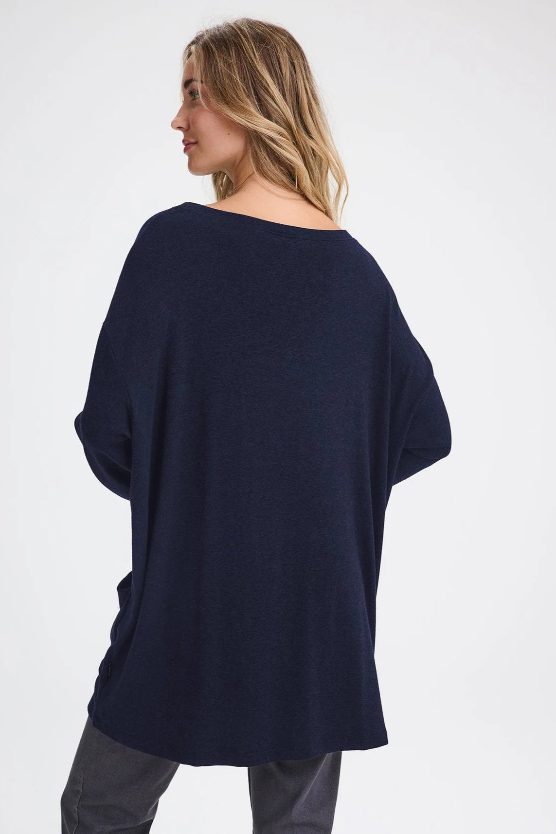 Back view of a woman wearing the Auclair Top by Message Factory in Navy, a bamboo/cotton/modal knit with a wide neck, long drop sleeves, a hi-low hem, and front patch pockets. She is wearing it with gray pants and standing in front of a white background.
