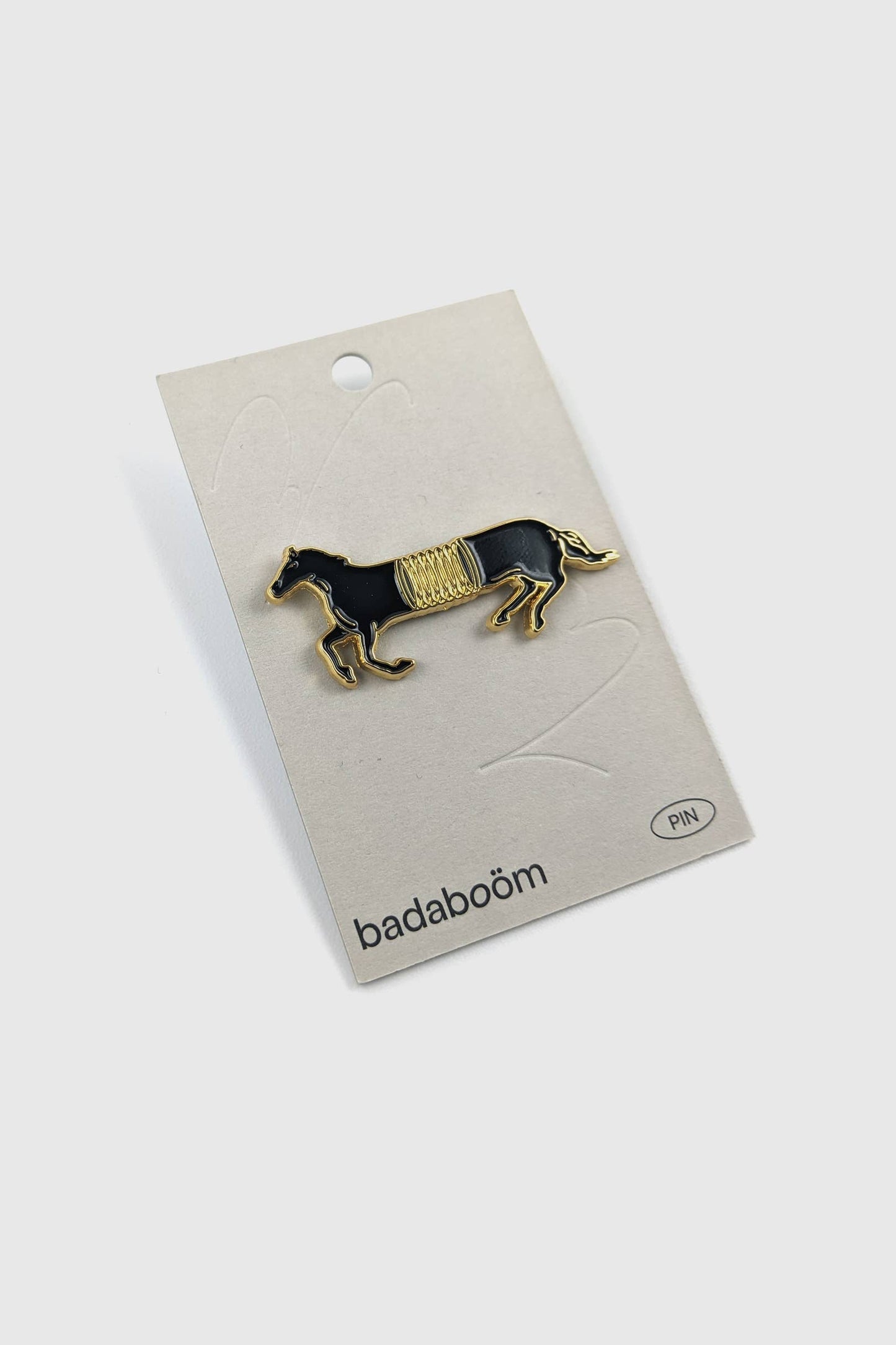 The Spring Horse Pin by Badaboöm Studio, featuring a black horse with a gold spring in the mid-section, is shown on a beige card against a white background.