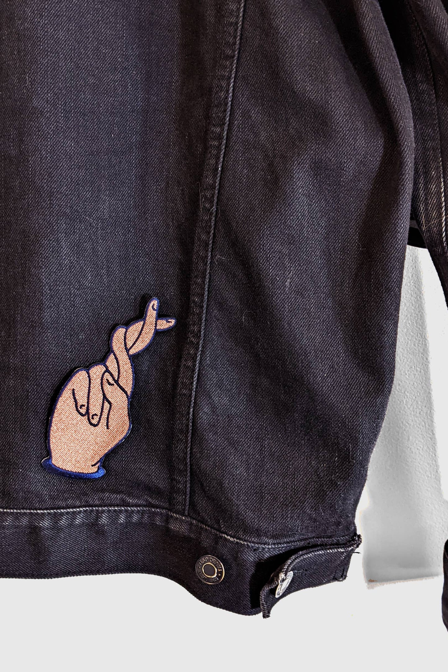 The Twist Patch by Badaboöm Studio, showing extra long crossed fingers, is shown on the back of a dark denim jacket.