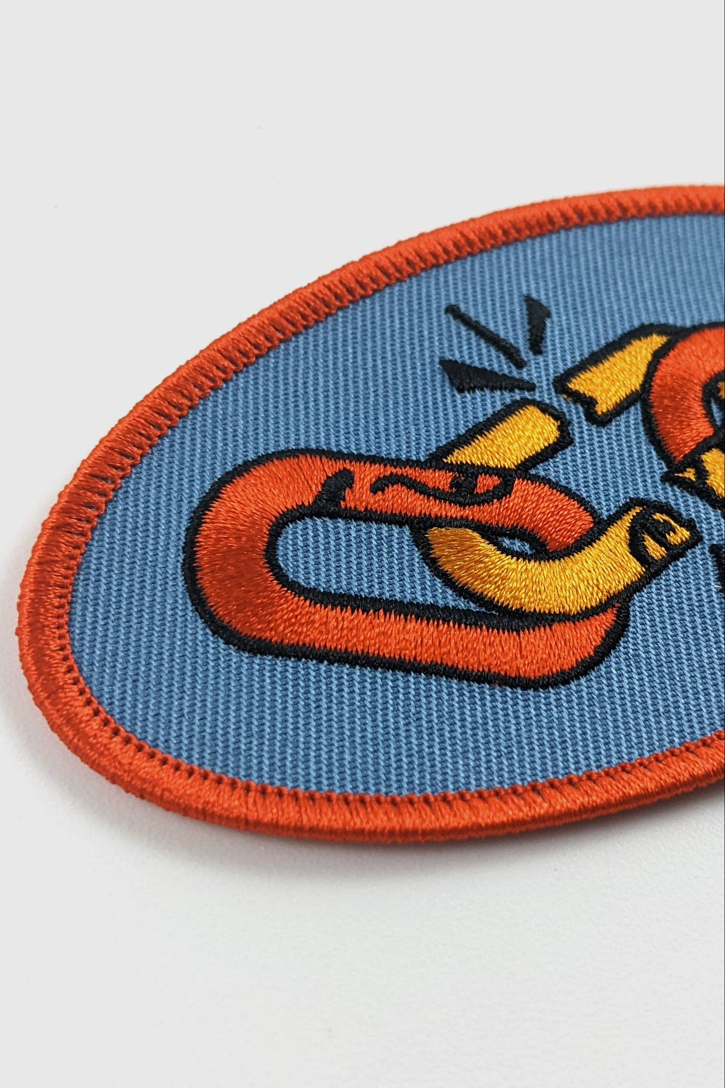 Embroidered Weakest Link patch from Badaboöm Studio, with a design of two multicoloured chain links on a blue background, bordered by orange
