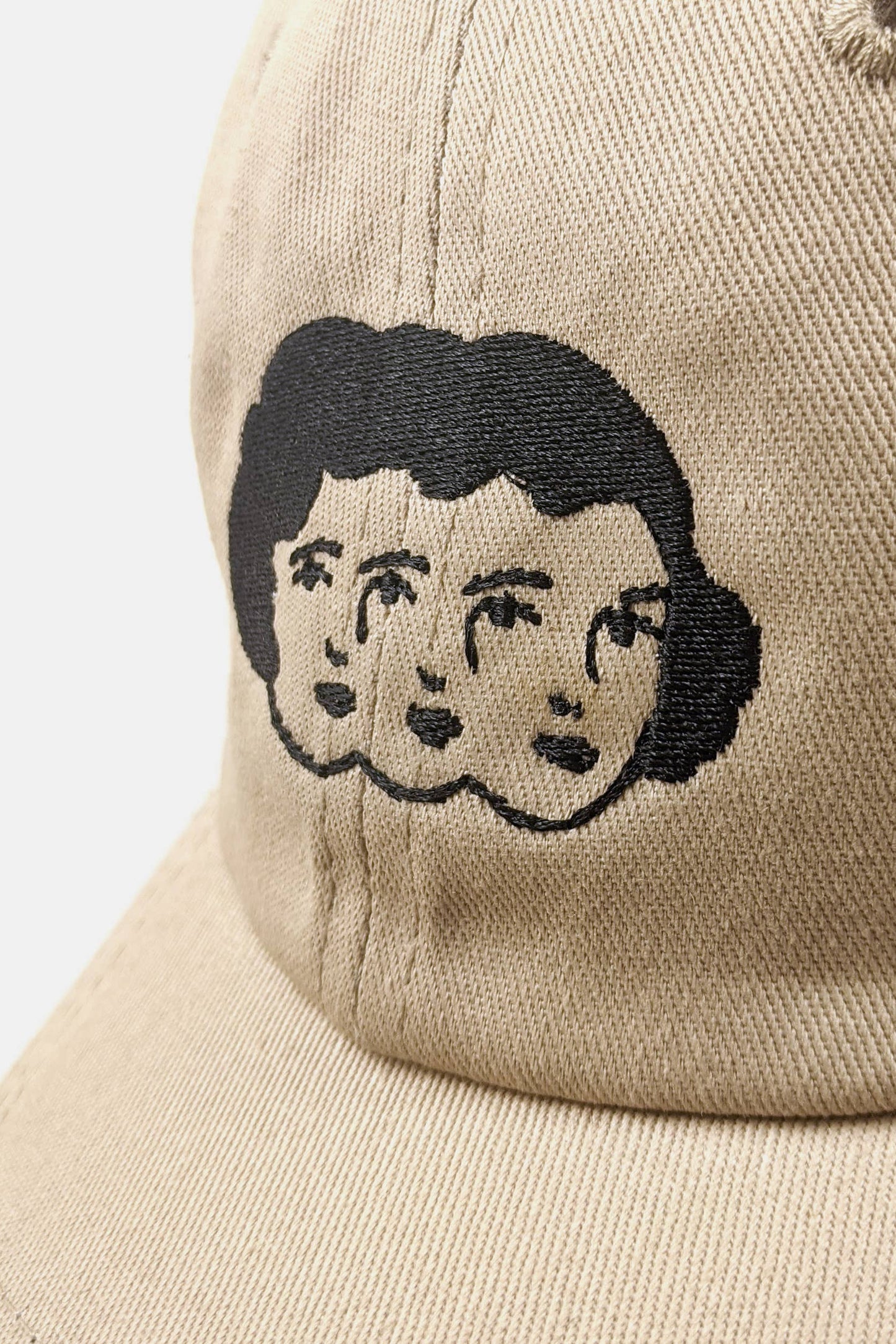 Close up view of the Triplets Hat from Badaboöm Studio, featuring embroidery of a woman's face in triplicate in black on a tan background, shown against a light grey background.