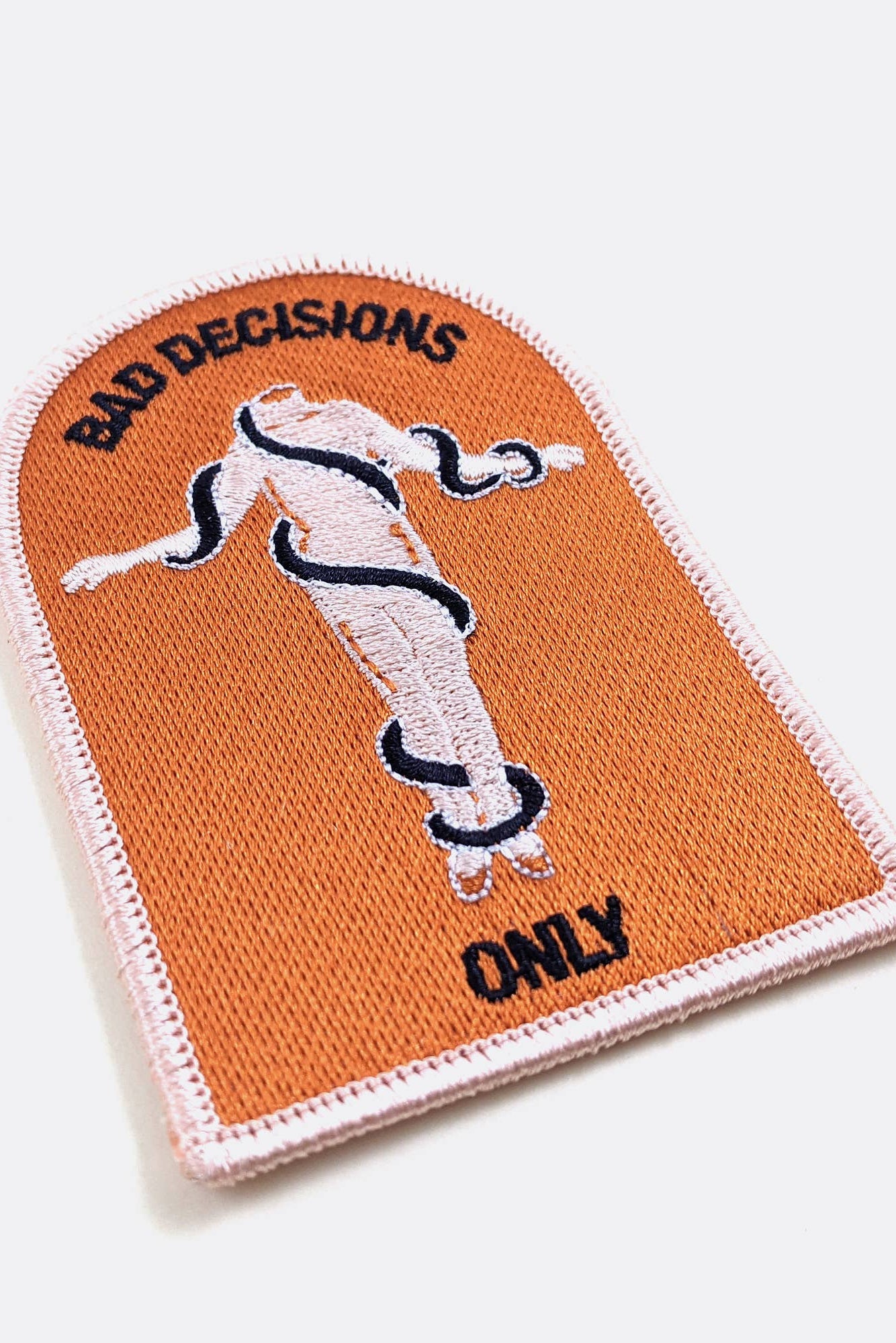 An arched embroidered patch from Badaboöm Studio with a peach-orange background, featuring a white female figure wrapped in a black snake, with the text 'BAD DECISIONS' at the top and 'ONLY' at the bottom.