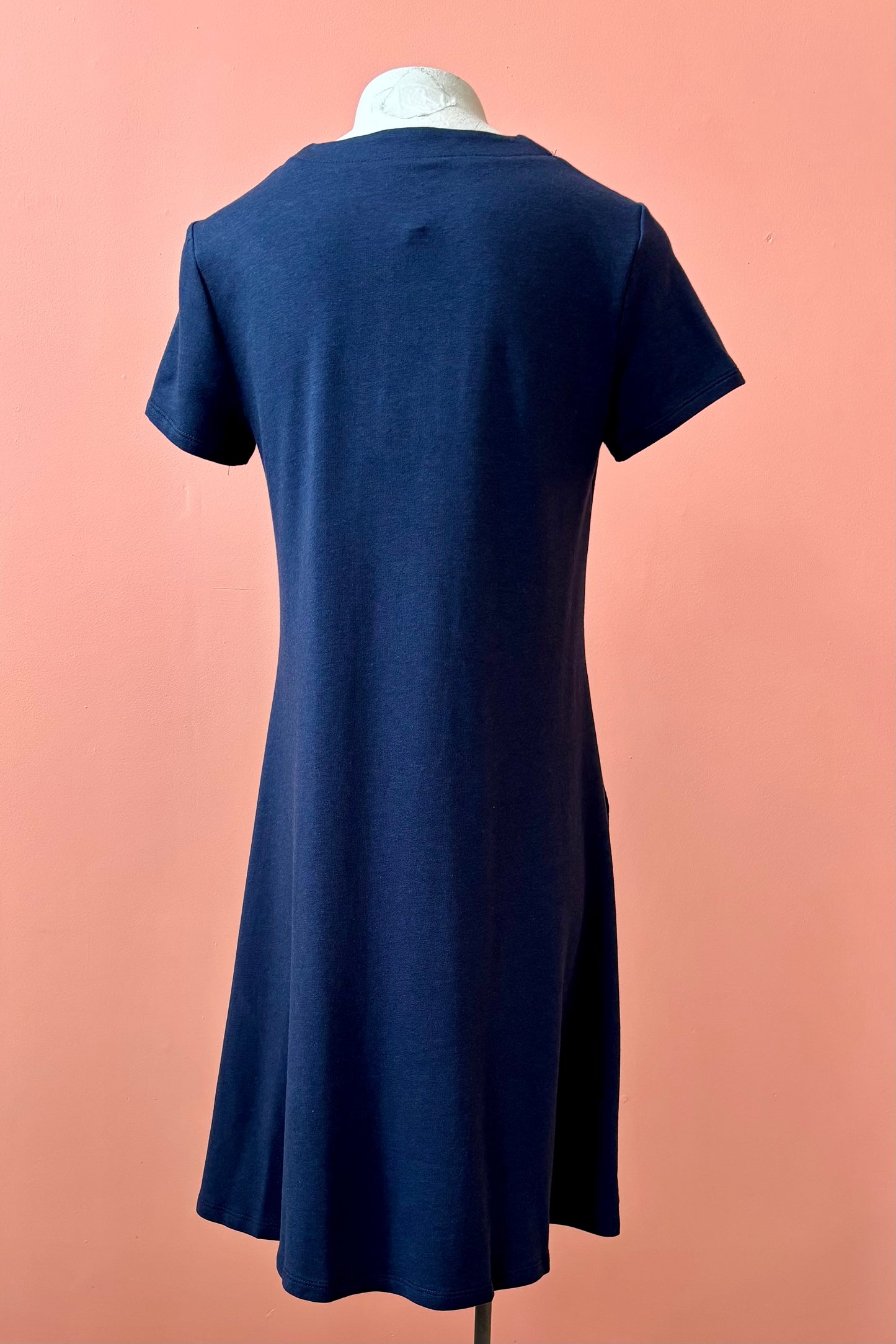 A back view of the Gaia Dress by Pure Essence in Navy, a short sleeved knee length dress with a V-neck, A-line shape, and pockets, is shown on a mannequin against a coral wall.
