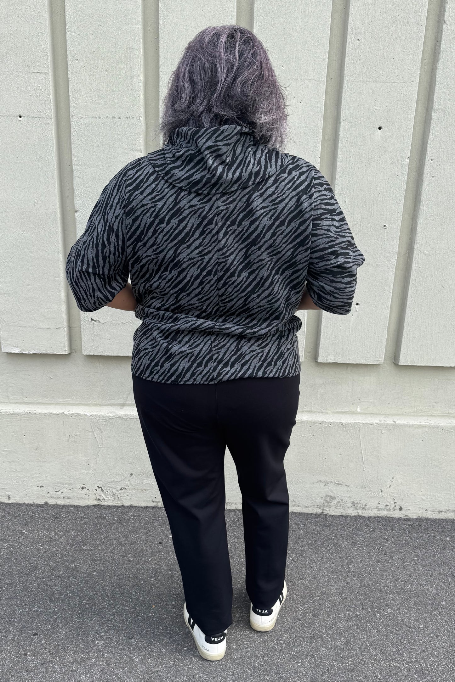Back view of a woman wearing the Francis Tunic by Pure in Black/Charcoal animal print, featuring a turtleneck, 3/4 sleeves and two patch pockets. She is wearing it with Black pants and standing in front of a grey wall.