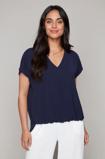 A woman wearing the Oakley Woven Top by Compli K in Navy, a short sleeved, loose fitting top with a V-neck. She's wearing it with white pants and sgtanding in front of a beige background.