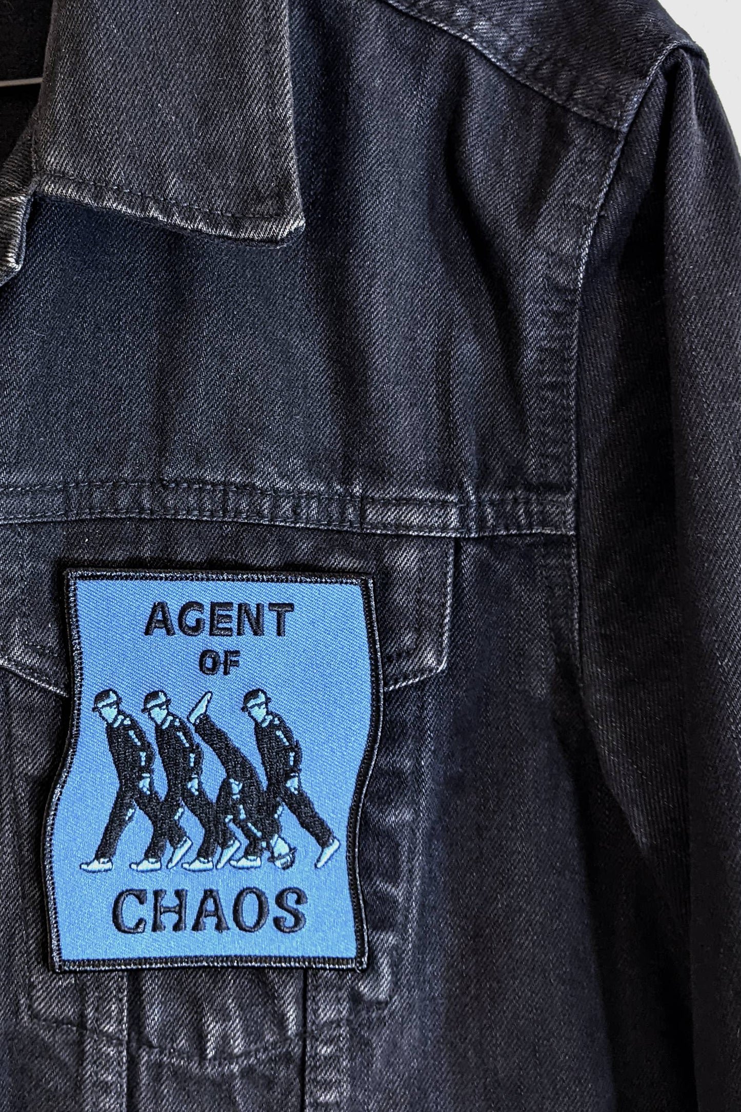 The Agent of Chaos patch from Badaboöm Studio, a rectangular blue patch depicting three men walking upright and one walking upside down, with the words of Agent of Chaos in black text, is shown on a denim jacket.