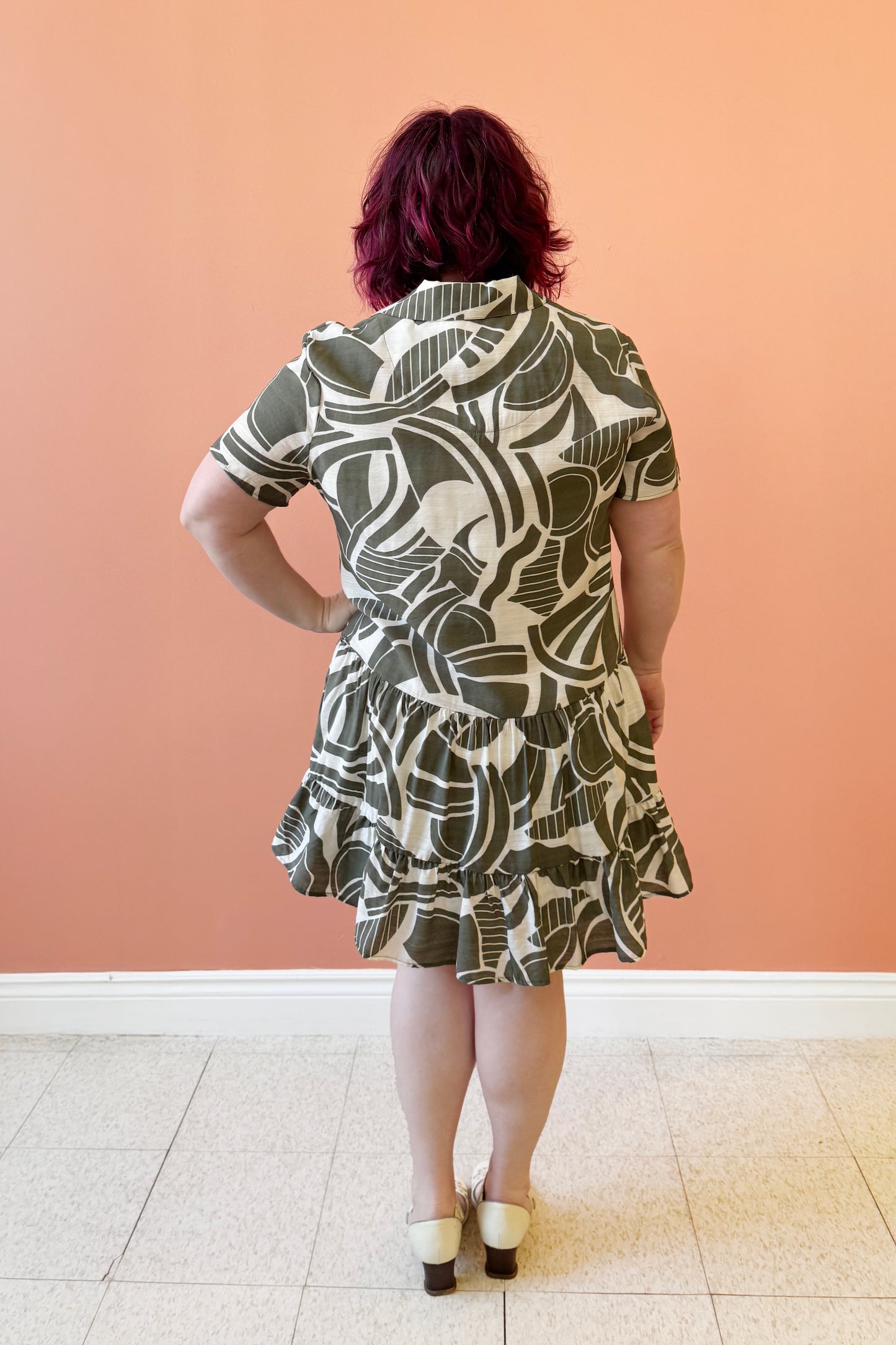 Back view of Allana wearing the Olympia Dress by Compli K in a Green/Cream geometric print, a short-sleeved dress with a blouse collar, button front, short tiered skirt with ruffles, and a slip dress under-layer. She is standing in front of a coral wall.