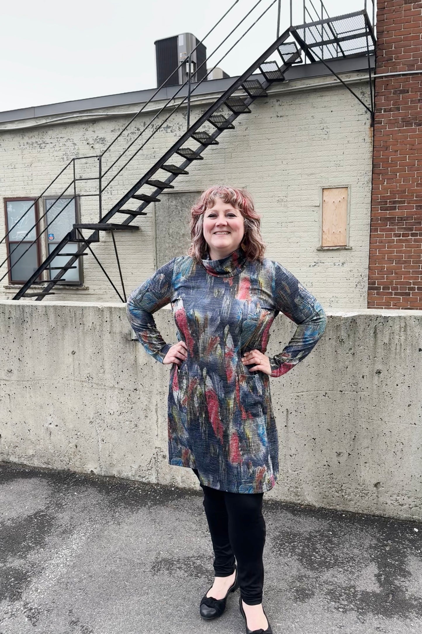 A woman wearing the Dakota Dress by Pure Essence, featuring a cowl neck, long sleeves, patch pockets, a knee-length hem, and a subtly sparkly Multicolour Print. She's wearing black leggings and standing outside in front of a brick building.