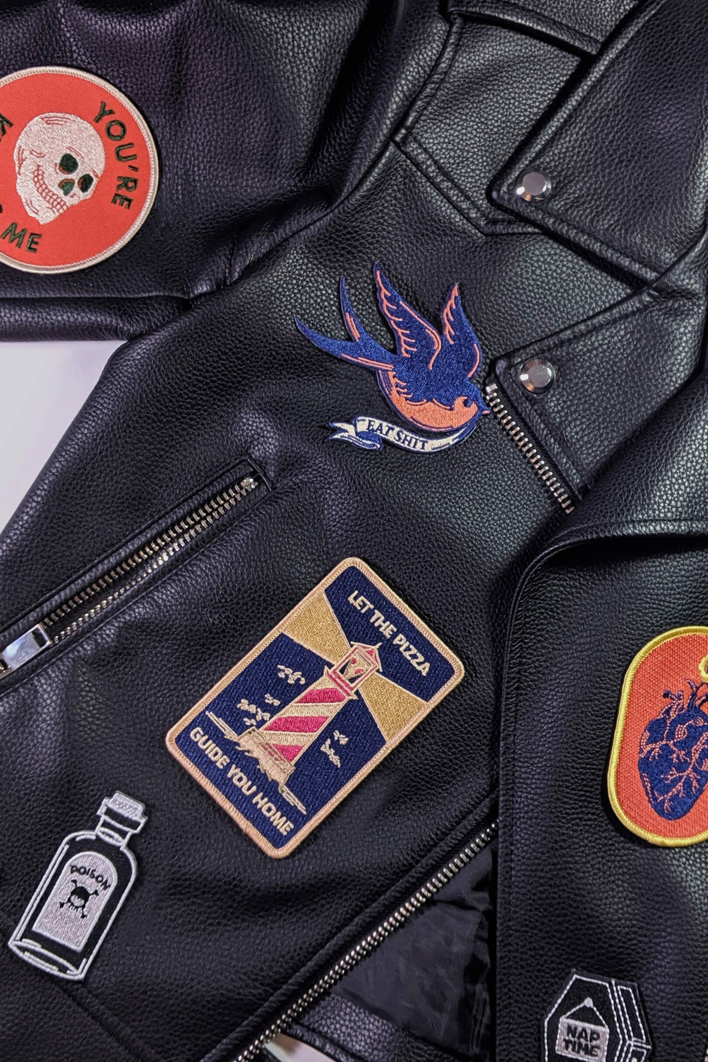 A vintage-style embroidered patch from Badaboöm Studio,
featuring a flying bird in navy blue and orange, with a banner reading 'EAT SHIT' below, shown on black leather jacket with other patches.