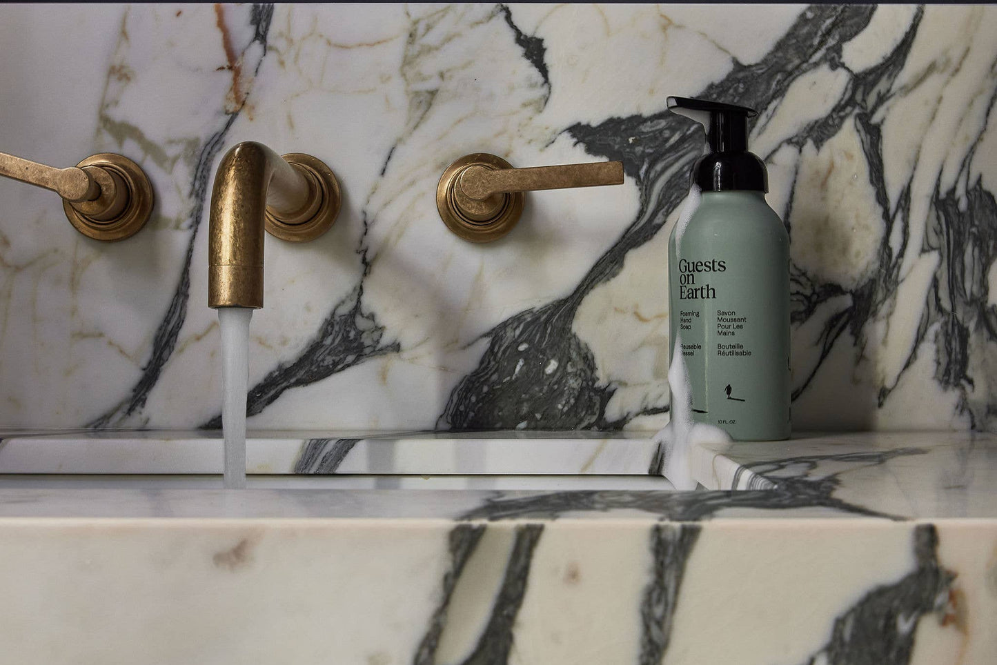 Marble bathroom sink with gold fixtures and a bottle of Guests on Earth foaming hand soap on a marble countertop.