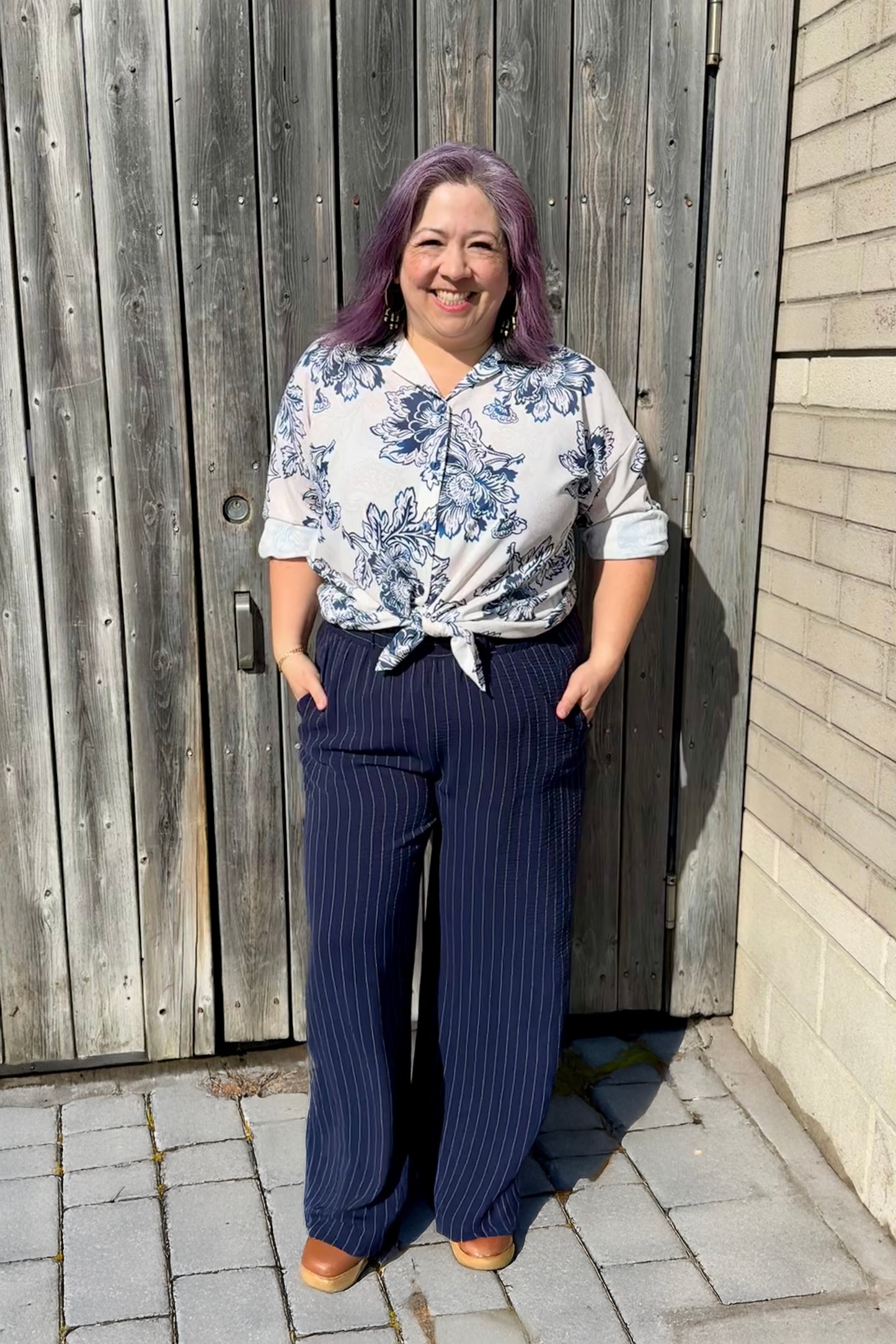 A woman wearing the Garnet Shirt by Pure Essence in Blue Stone floral print, a classic button front shirt with dropped shoulders. She is wearing with the Gabby Pants and standing in front of wooden fence.