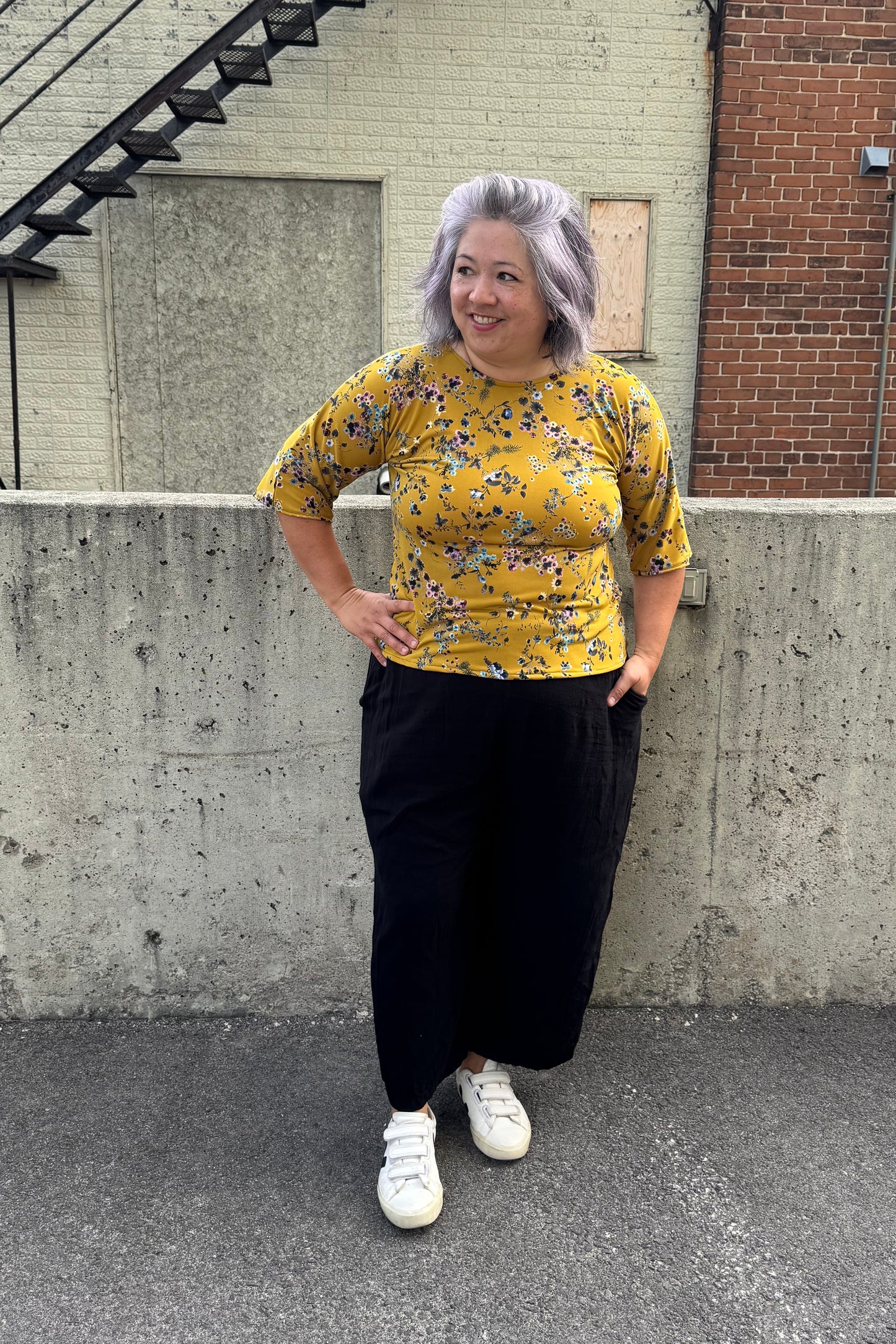 A woman wearing the Oversized Top by SI Design in Yellow Floral, featuring a scoop neck, elbow-length sleeves, and a slightly relaxed fit. She is wearing with black pants and standing outside in front of a brick building.