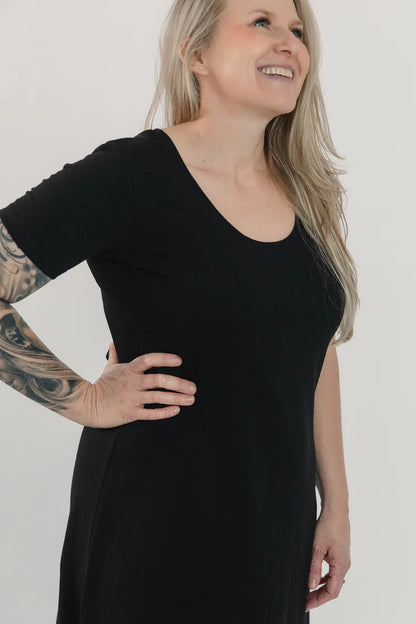 Side close up view of a woman wearing the Field Dress by Blondie in Black, with short sleeves, a scooped neck, and a contoured hem that hits at mid-calf and cuts up at the sides. She is standing in front of a white background. 