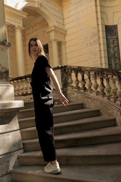 Side view of a woman wearing the Oslo Jumpsuit by MAS in Pepper, with round neck, short sleeves, adjustable waist tie, and wide legs. She is walking down a set out of outdoor stairs. 