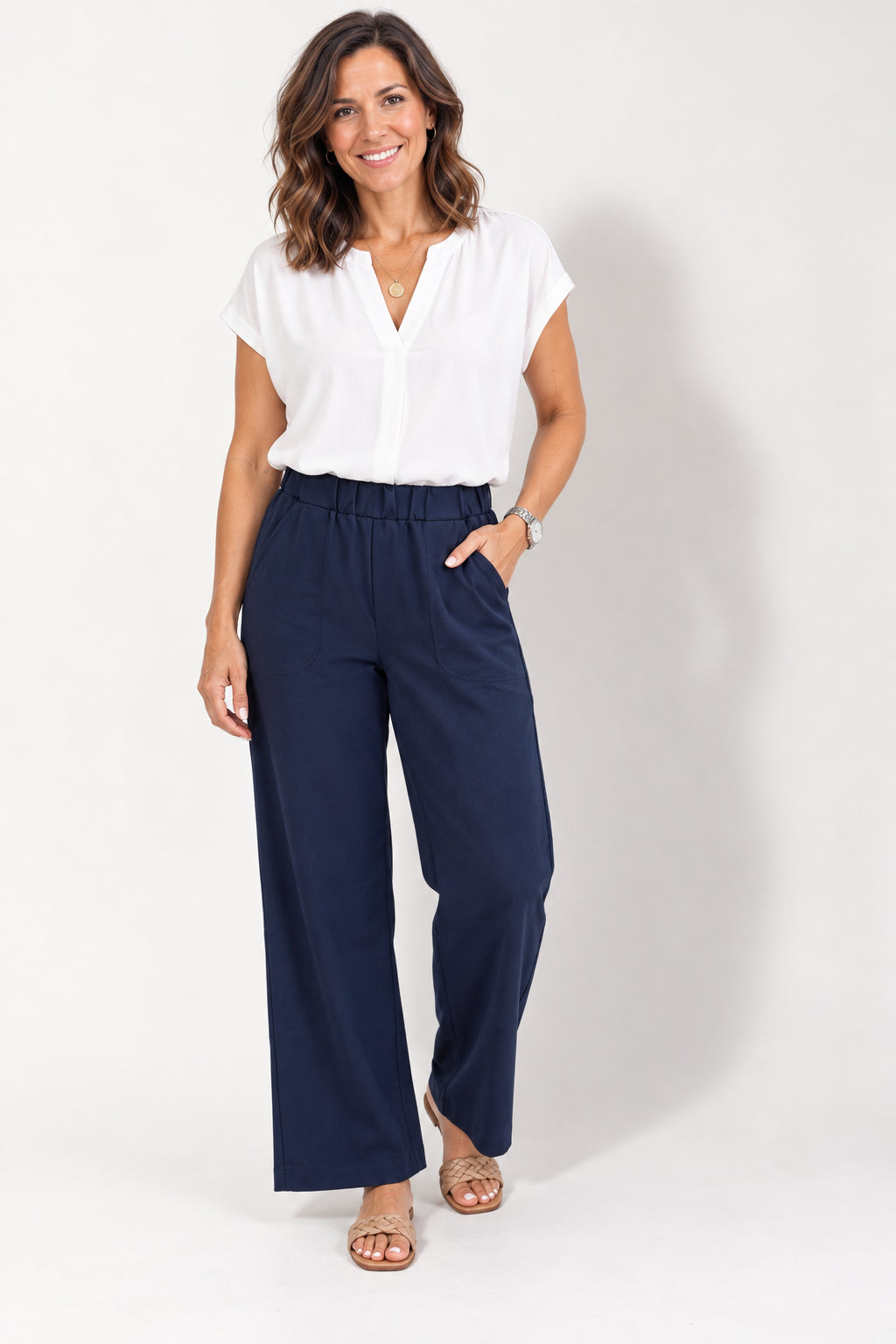A woman wearing the Luisa Ponte Pants by Tangente in Navy, pull-on pants with an elastic waist, front pleats, and side pockets. She is wearing them with a white top and standing in front of a white studio background. 