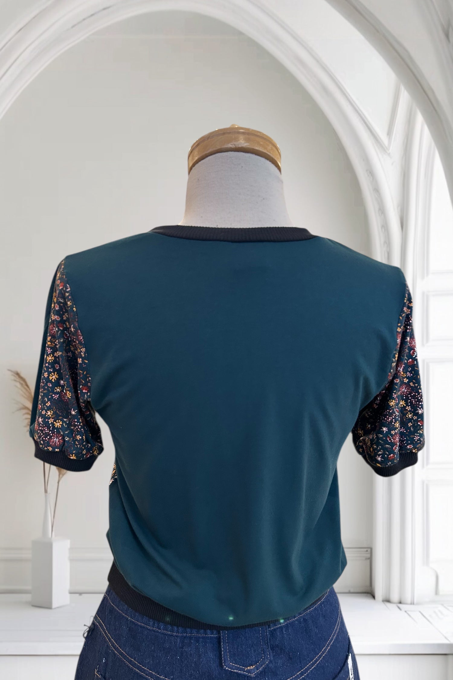 A back view of the Darling T-Shirt by Slak in Floral with Green, a short sleeved, semi-fitted sweater with contrasting fabric at the cuffs, collar and hem, inserts on the sleeves, and a solid back, is shown on a mannequin against a white background. 