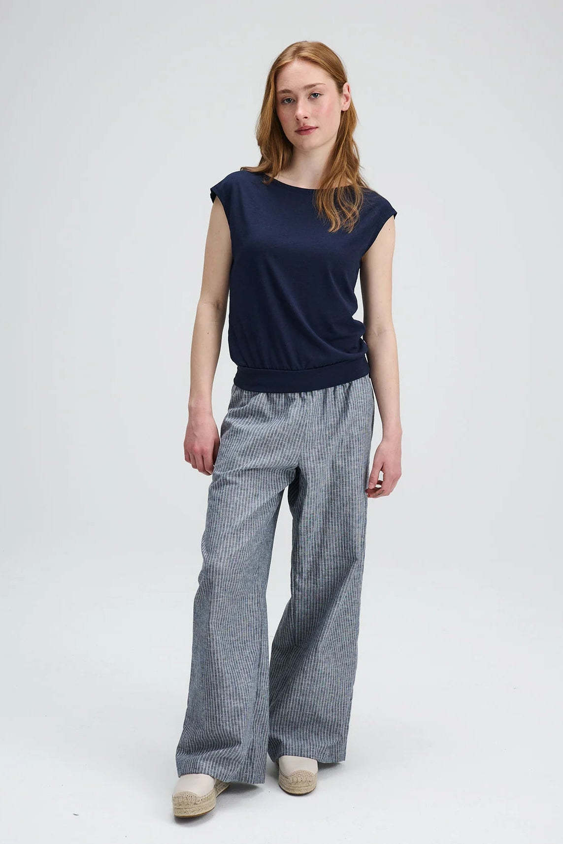 A woman wearing the Batiscan Top by Message Factory in Navy, with short dropped sleeves and a wide neckline, plus a band at the hem. She is wearing it with the Liberte pants in Grey Stripe and standing against a light grey background  
