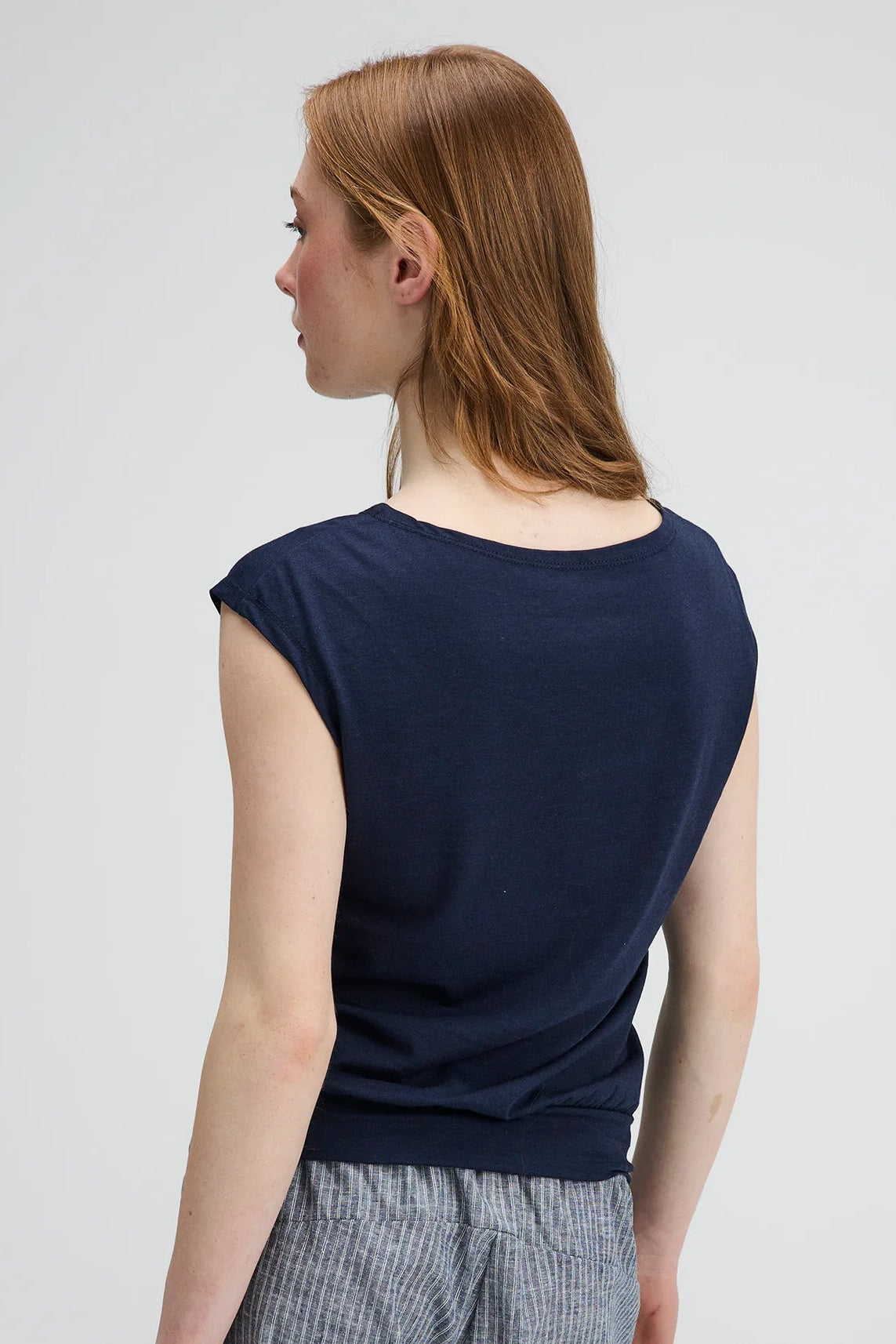Back waist up view of a woman wearing the Batiscan Top by Message Factory in Navy, with short dropped sleeves and a wide neckline, plus a band at the hem. She is wearing it with the Liberte pants in Grey Stripe and standing against a light grey background  