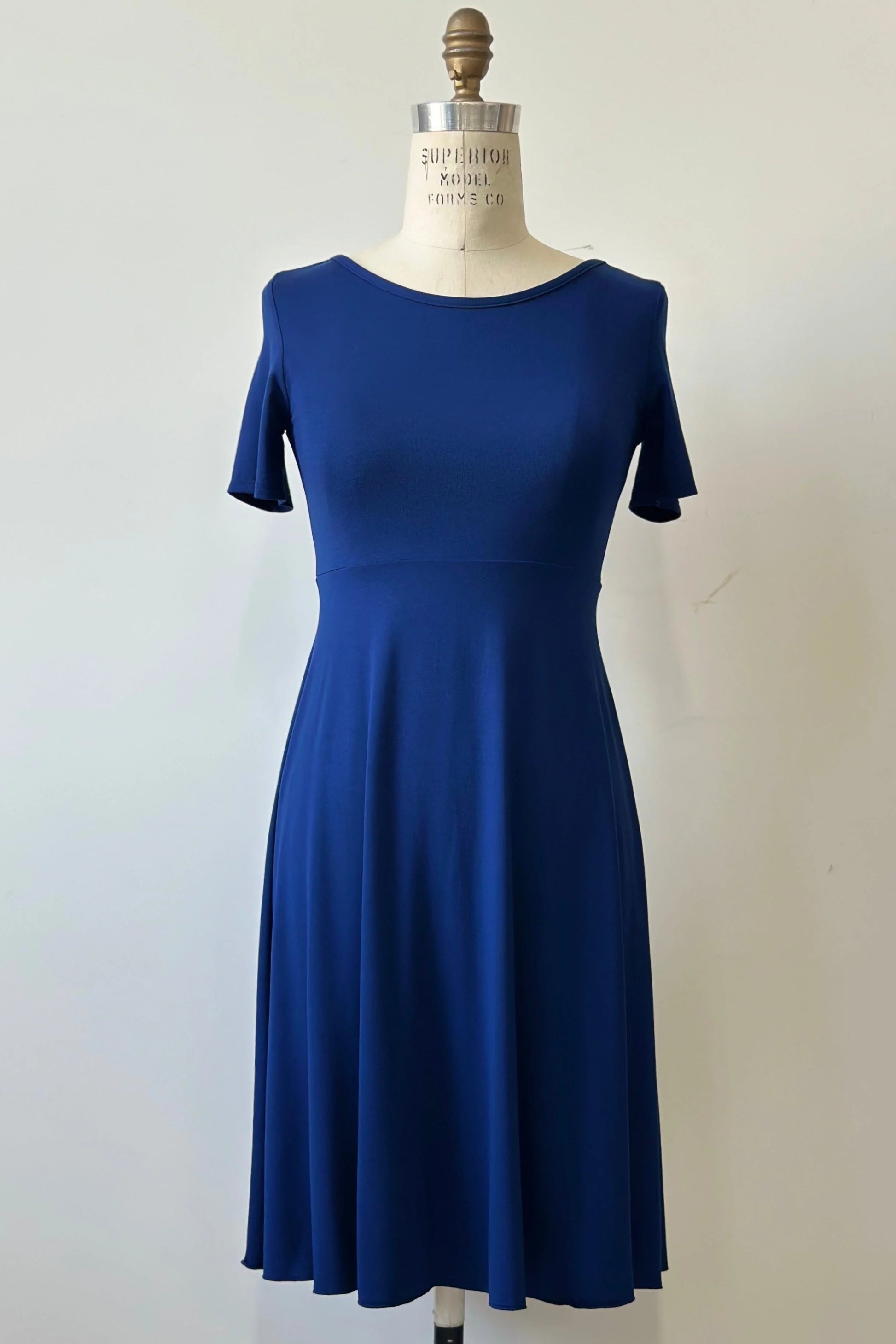 The Luma dress by Kollantai in Admiral blue, featuring short sleeves, a wide neck, an empire waist, and a full flared skirt that falls to the knee, is shown on a mannequin against a white background.