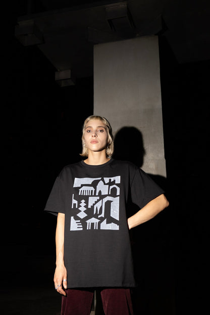 A woman wearing the Tiger's Eye Screen-printed T-shirt by Eve Lavoie, an oversized black t-shirt with a white graphic print on the front. She is wearing it with burgundy pants and standing in a dark industrial setting. 