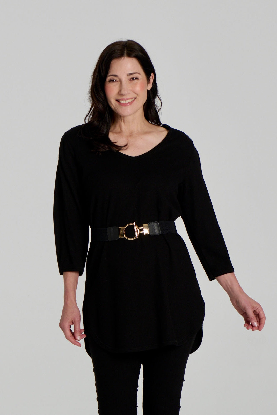  A woman wearing the Clara Tunic by Kollontai in Black, a stretch corduroy piece with a V-neck, a rounded hem, 3/4 sleeves, and inverted pleats at the shoulders. She is wearing it belted over black leggings and standing against a grey background. 