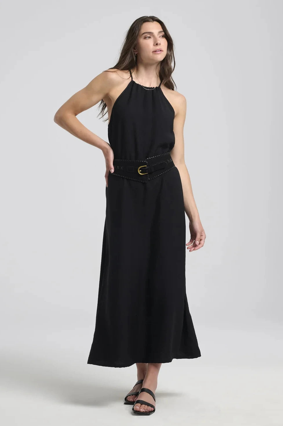 A woman wearing the Anwen Dress by Kollontai in Black, a loose-fitting rayon maxi dress with an adjustable halter neckline. She is wearing it with a belt and is standing in front of a white background. 