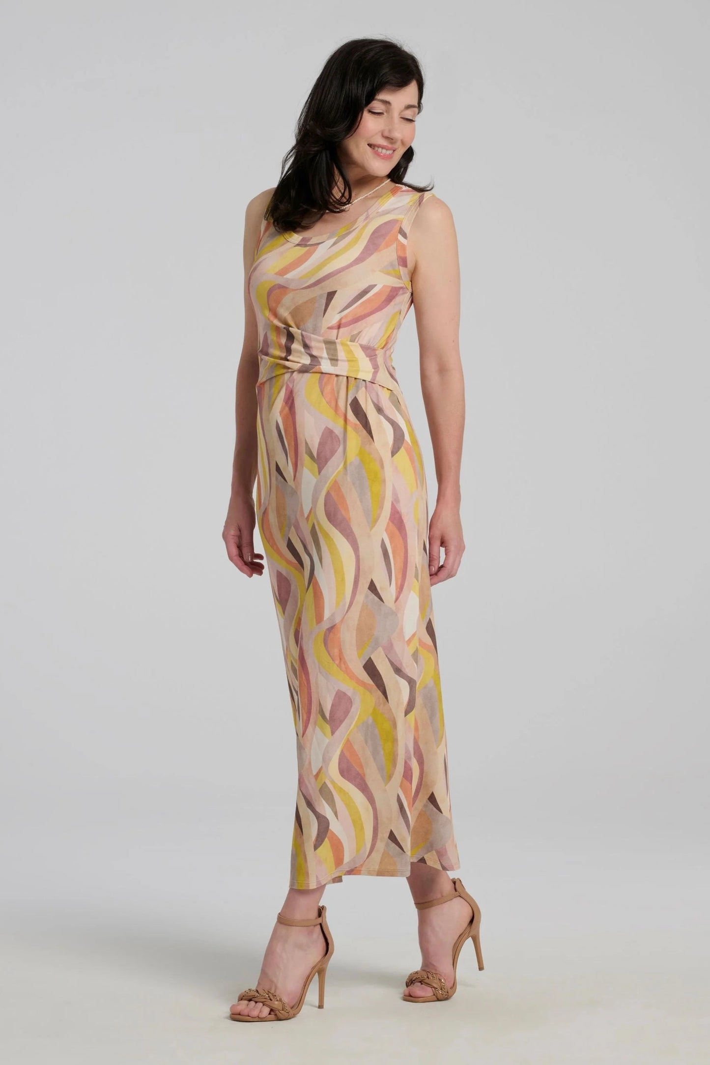 A woman wearing the Serina Dress by Kollontai in Peach print, a long, slim cut sleeveless dress with a soft pleat at the waist. She is standing in front of a white background. 