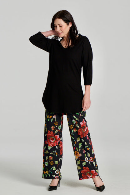 A woman wearing the Clara Tunic by Kollontai in Black, a stretch corduroy piece with a V-neck, a rounded hem, 3/4 sleeves, and inverted pleats at the shoulders. She is wearing it with the Elma Pants in floral and standing in front of a grey background.