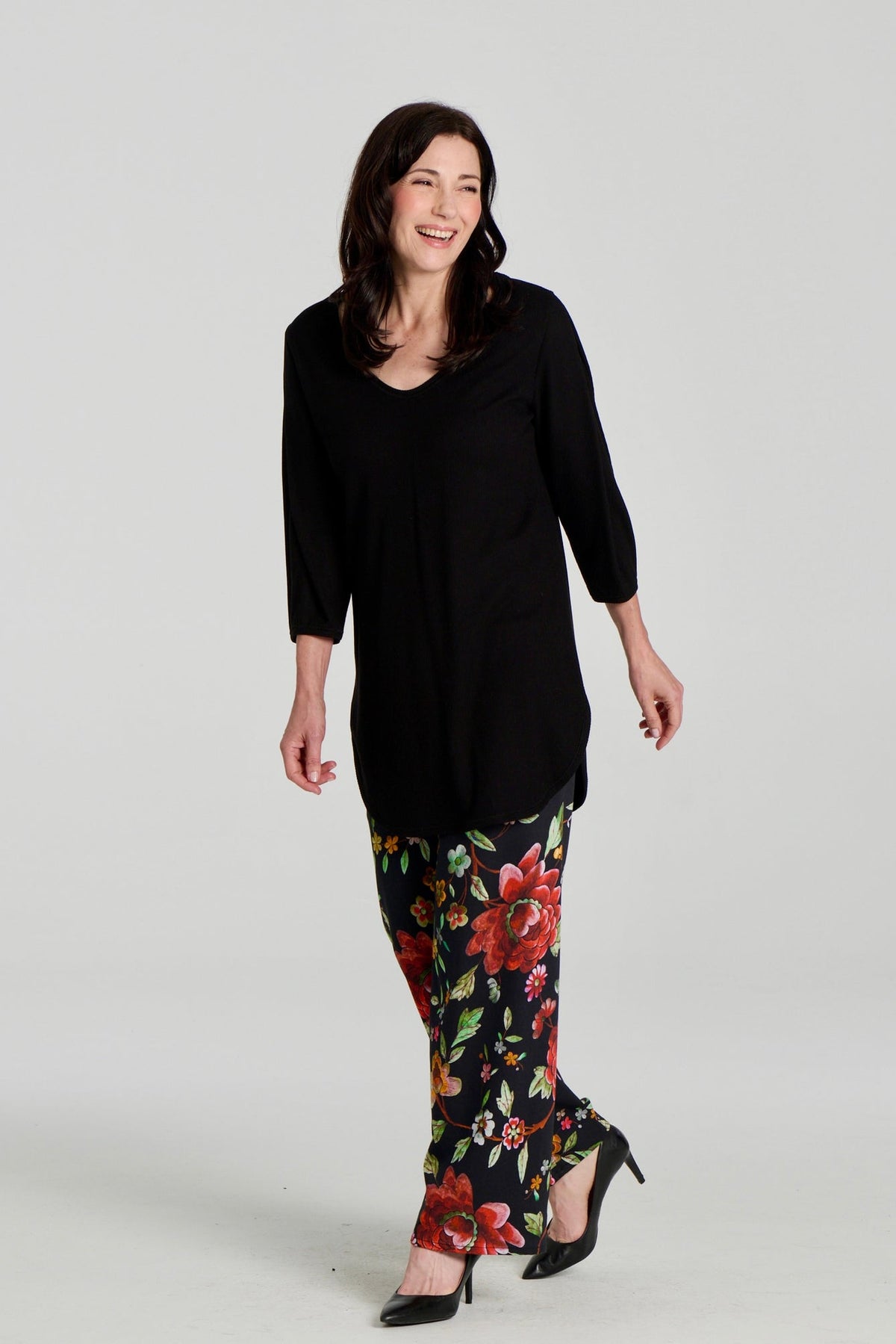 A woman wearing the Clara Tunic by Kollontai in Black, a stretch corduroy piece with a V-neck, a rounded hem, 3/4 sleeves, and inverted pleats at the shoulders. She is wearing it with the Elma Pants in floral and standing in front of a grey background.