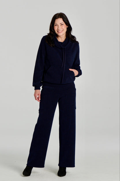 A woman wearing the Lorenzo Pants by Kollontai with the Ramos Sweater, both in a ribbed navy corduroy-like fabric. The Sweater has a large cowl neck with a drawstring and a kangaroo pocket, the pants have a wide pull on waist, straight legs, and large patch pockets. She is standing against a white background. 