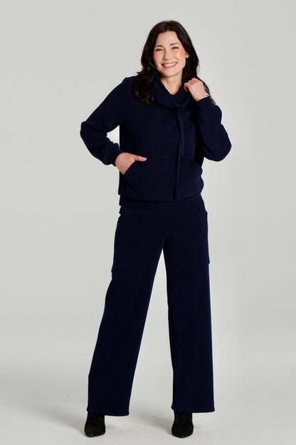 A woman wearing the Lorenzo Pants by Kollontai with the Ramos Sweater, both in a ribbed navy corduroy-like fabric. The Sweater has a large cowl neck with a drawstring and a kangaroo pocket, the pants have a wide pull on waist, straight legs, and large patch pockets. She is standing against a white background. 