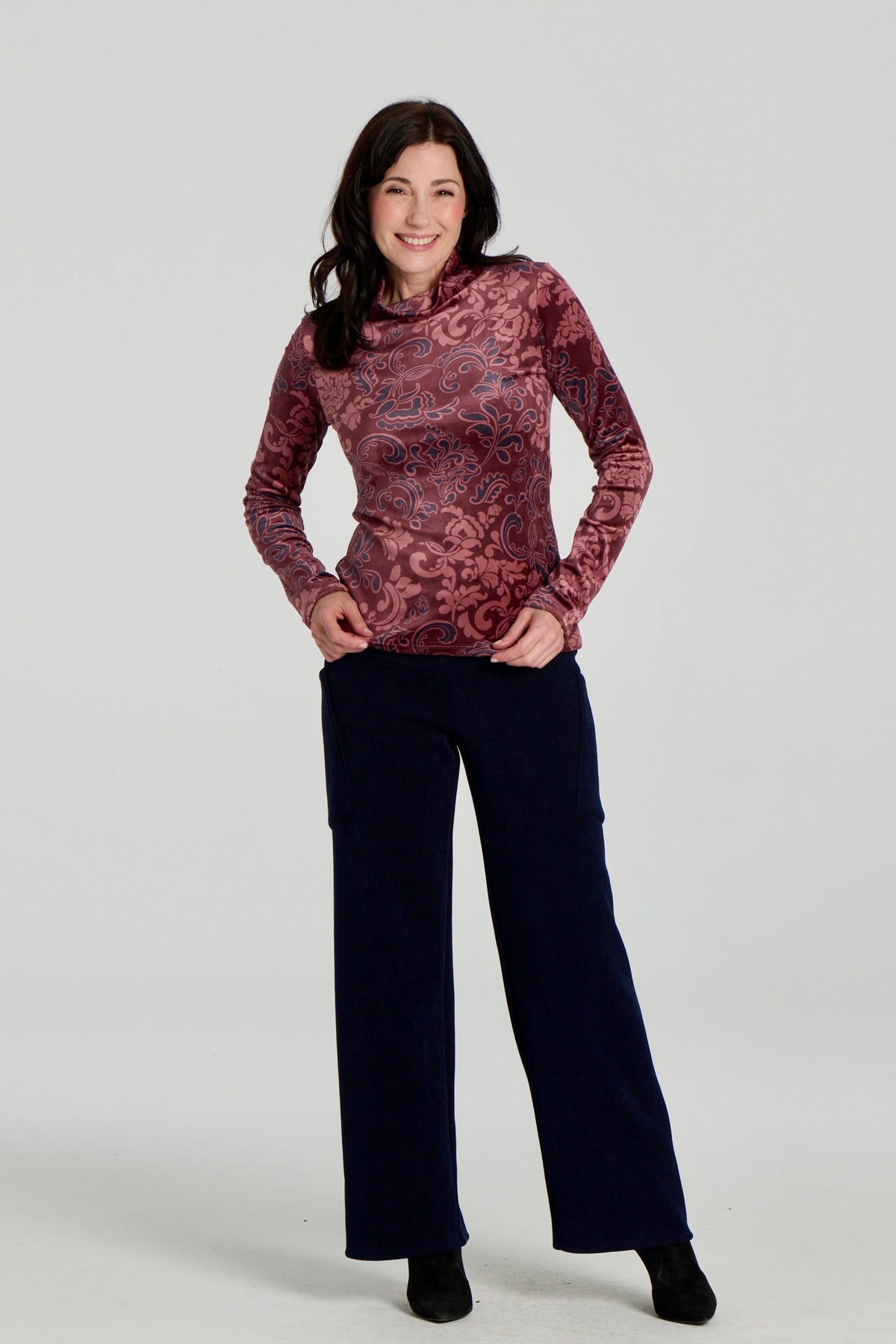 A woman wearing the Lorenzo Pants by Kollontai in a ribbed navy corduroy-like fabric, with a wide pull on waist, straight legs, and large patch pockets. She is wearing them with the Everdeen Sweater and standing against a white background. 