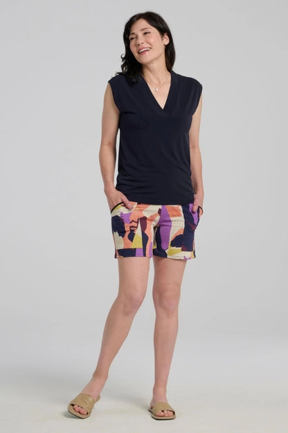 A woman wearing the Vela Shorts by Kollontai in Multicolour abstract print, with a pull-on waist, straight cut, and patch pockets. She is wearing them with  dark sleeveless top against a plain background