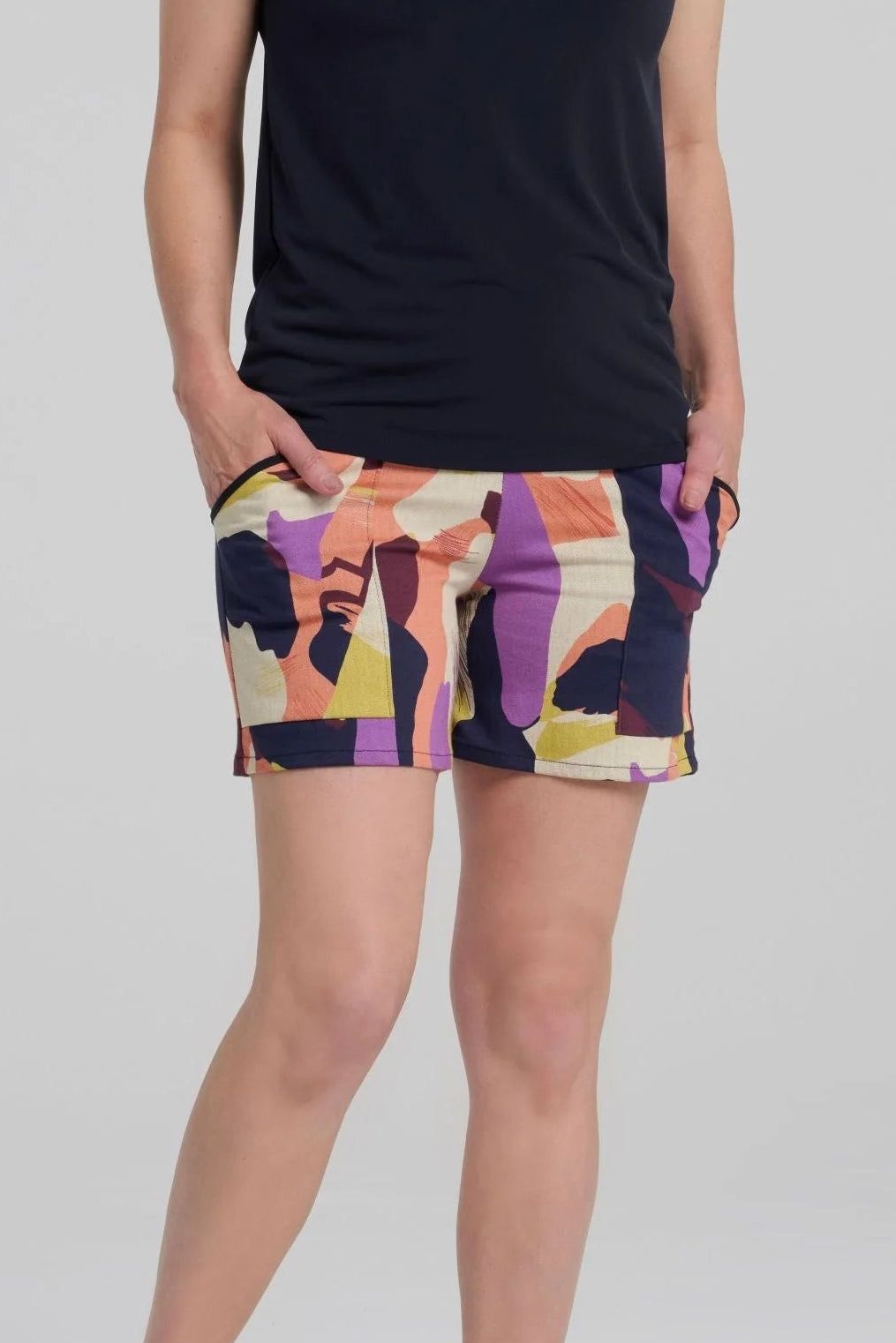 Torso view of a woman wearing the Vela Shorts by Kollontai in Multicolour abstract print, with a pull-on waist, straight cut, and patch pockets. She is wearing them with  dark sleeveless top against a plain background