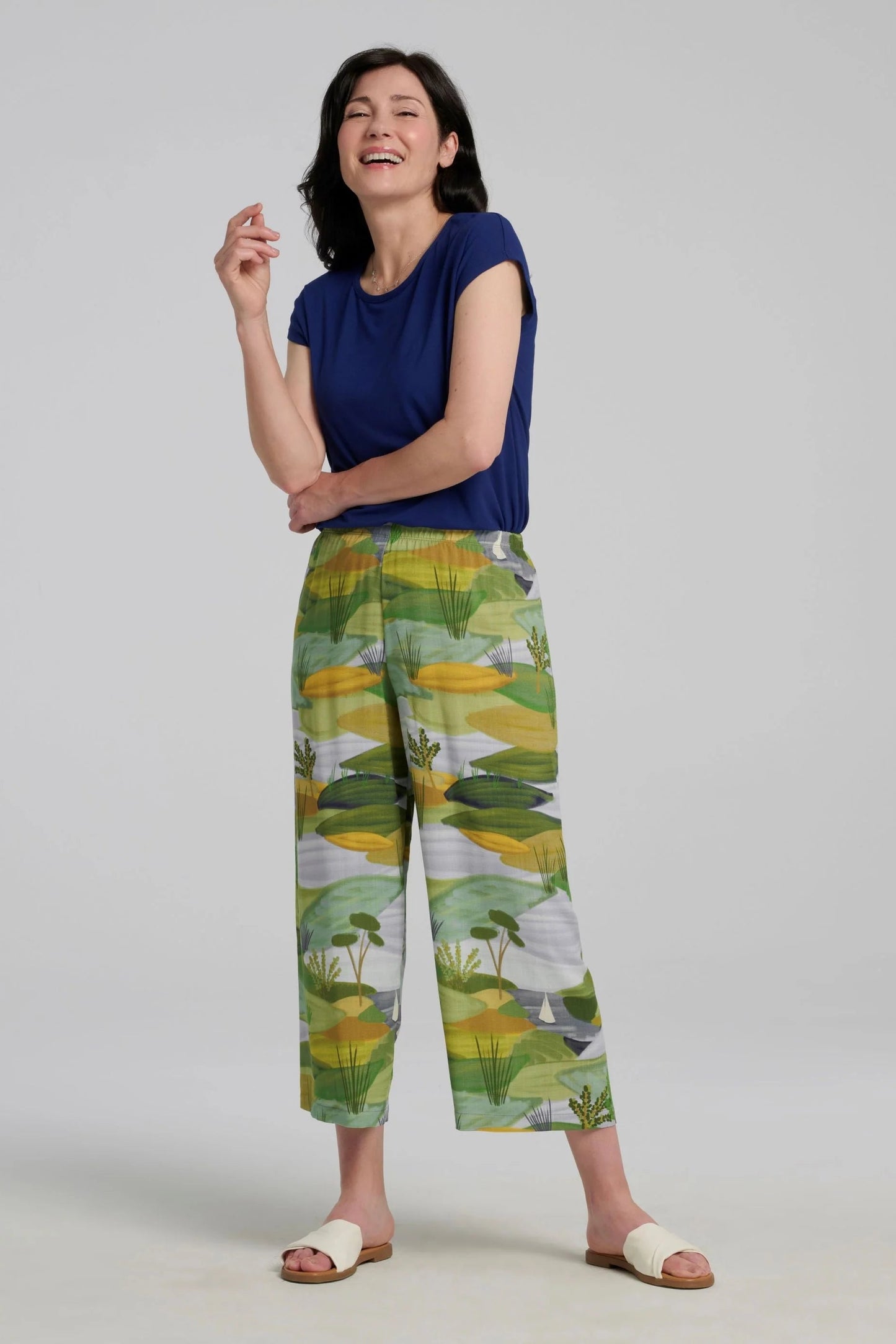 A woman wearing the Paraiso Pants by Kollontai in a Granny Smith toned nature print, with an elastic waist and wide cropped legs with side slits. She is wearing them with a blue  top and standing in front of a plain background