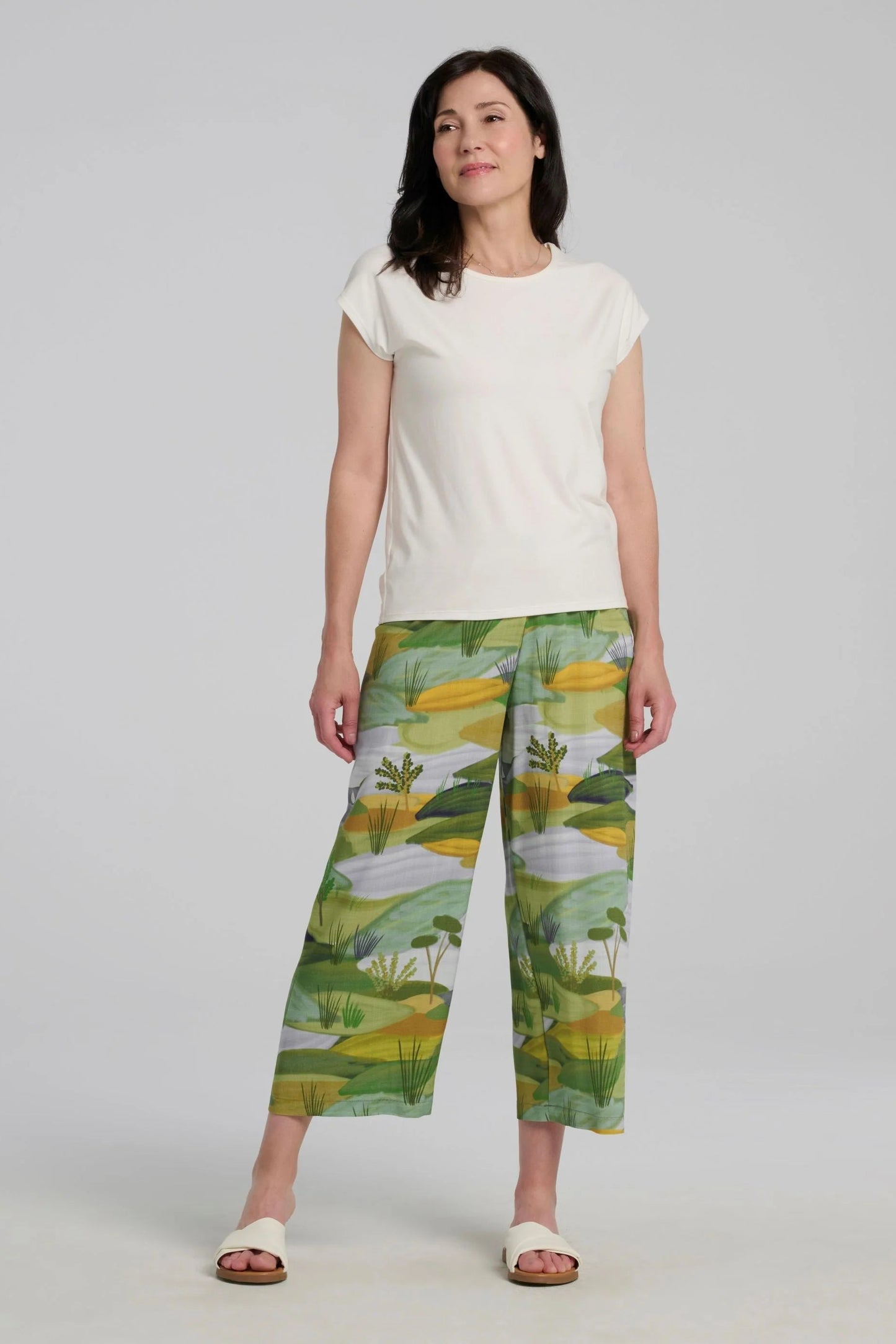 A woman wearing the Paraiso Pants by Kollontai in a Granny Smith toned nature print, with an elastic waist and wide cropped legs with side slits. She is wearing them with the Eowyn Top in off-white and standing in front of a plain background