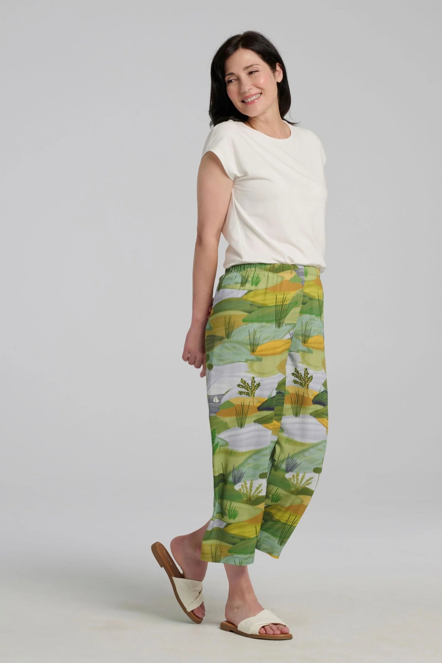 A woman wearing the Paraiso Pants by Kollontai in a Granny Smith toned nature print, with an elastic waist and wide cropped legs with side slits. She is wearing them with the Eowyn Top in off-white and standing in front of a plain background