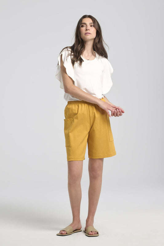 A woman wearing the Morgana T-Shirt by Kollontai in White, a loose fitting top with a round neck and fly-away sleeves that fall to the elbow. She is wearing it with yellow shorts and standing in front of a white background. 