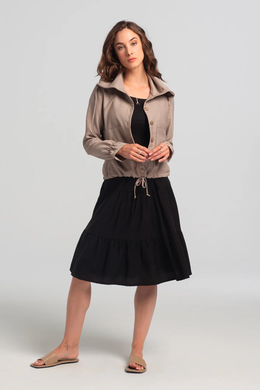 A woman wearing the Al-Juzza Skirt by Kollontai in Black with a beige jacket, standing in front of a white background 