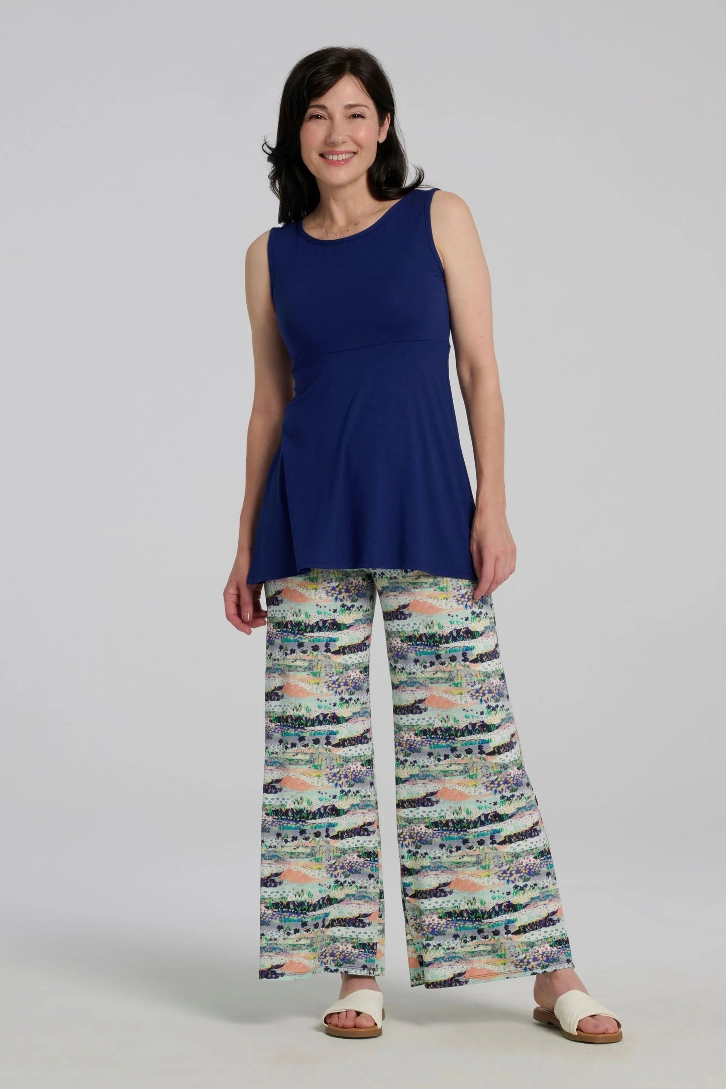 A woman wearing the Nira Top by Kollontai in Admiral blue, a sleeveless top with a wide neck, an empire waist, and a cut that's fitted through the bodice and flares away from the torso. She is wearing it with colourful patterned pants and standing against a grey background.