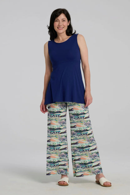 A woman wearing the Nira Top by Kollontai in Admiral blue, a sleeveless top with a wide neck, an empire waist, and a cut that's fitted through the bodice and flares away from the torso. She is wearing it with colourful patterned pants and standing against a grey background.