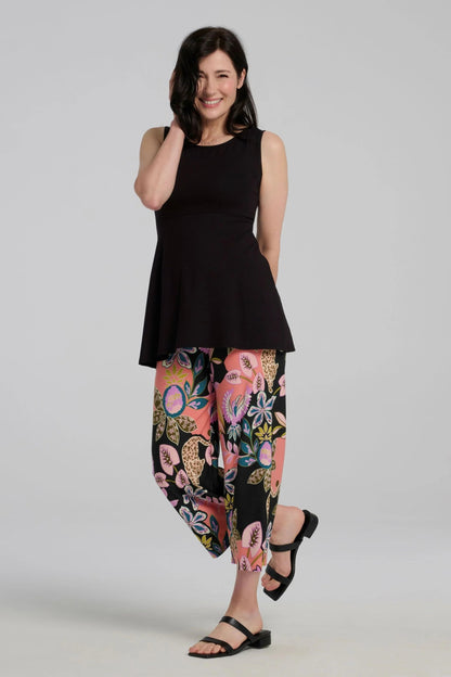 A woman wearing the Nira Top by Kollontai in Black, a sleeveless top with a wide neck, an empire waist, and a cut that's fitted through the bodice and flares away from the torso. She is wearing it with colourful patterned pants and standing against a grey background.