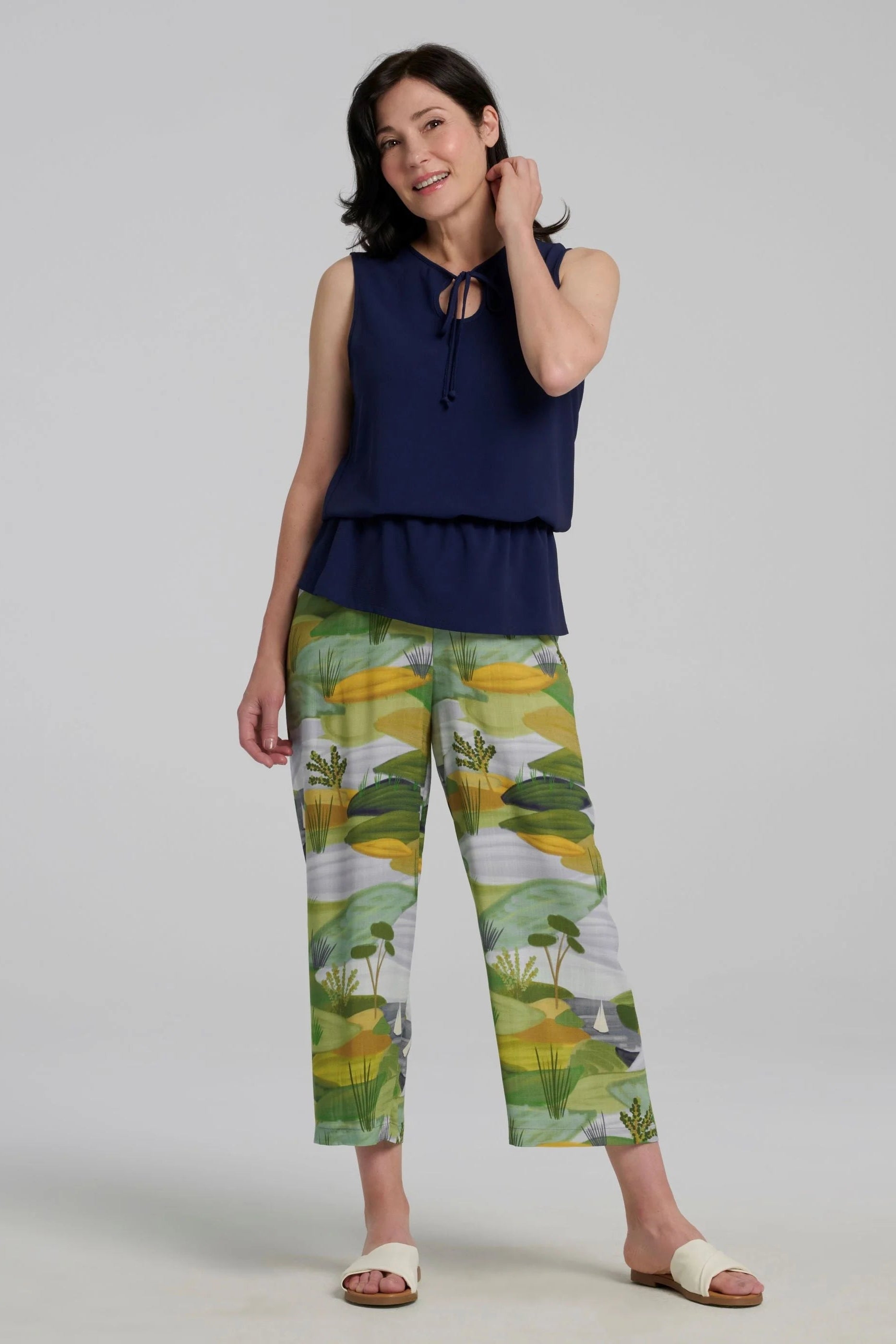 A woman wearing the Paraiso Pants by Kollontai in a Granny Smith toned nature print, with an elastic waist and wide cropped legs with side slits. She is wearing them with a blue top and standing in front of a plain background