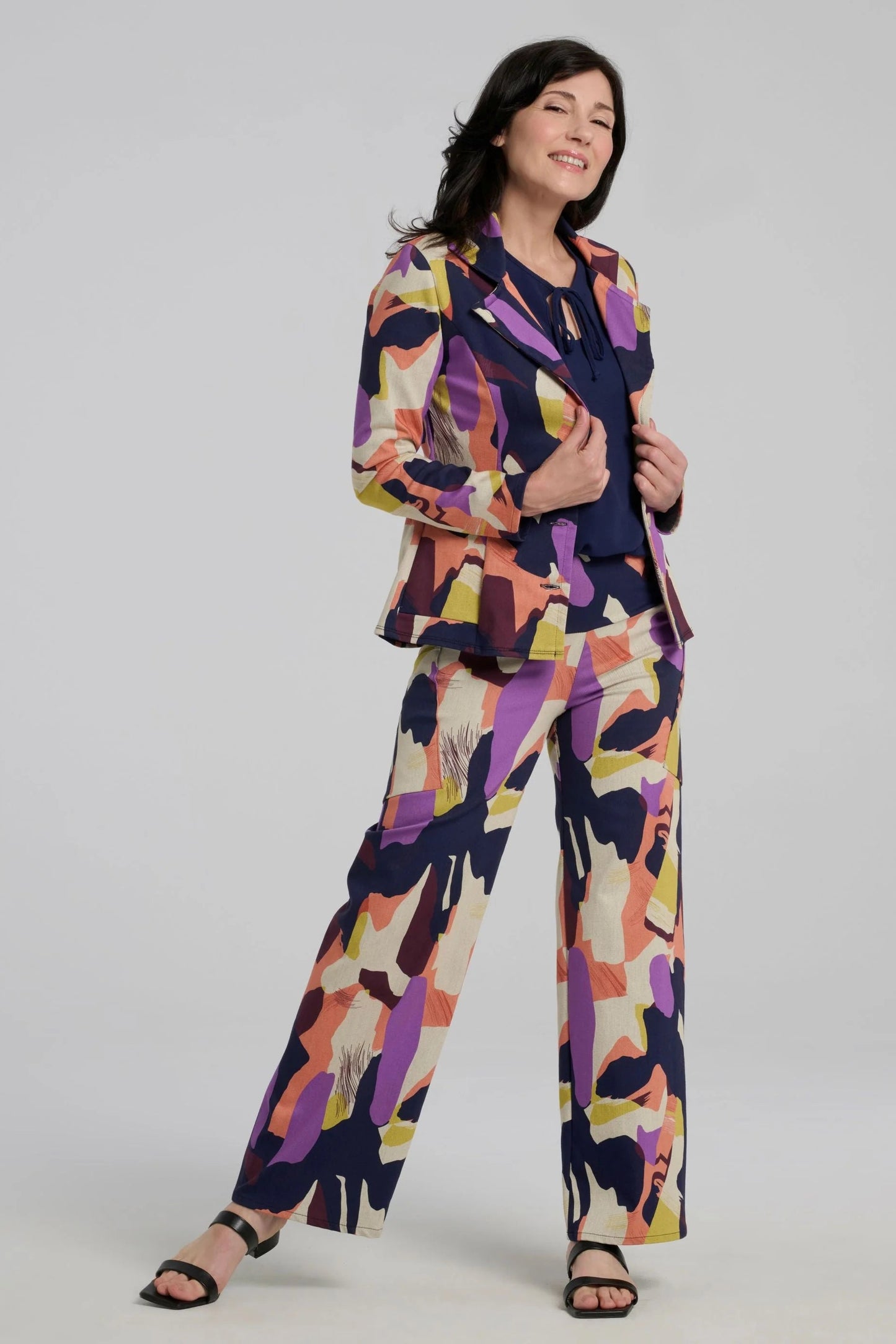 A woman wearing the Elma Pants by Kollontai in a Multicolour abstract print, with a wide pull-one waistband, straight legs, and slanted pockets. She is wearing them with a navy top matching jacket and standing in front of a plain background