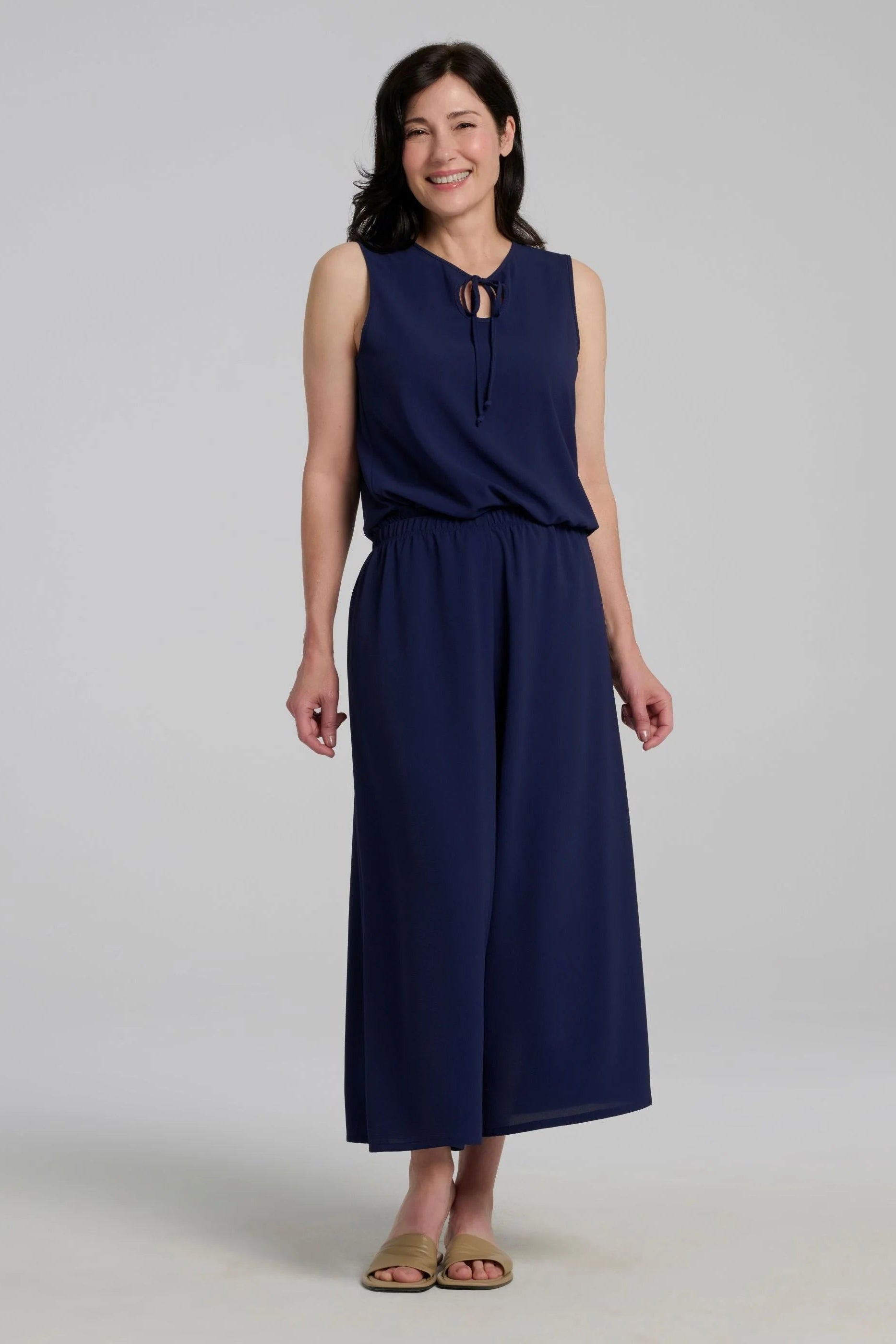 A woman wearing the Serena Top from Kollontai in Navy, a sleeveless top with a keyhole and tie neckline, elastic waist and flared peplum hem. She is wearing it with the Ishara Pants against a a grey background