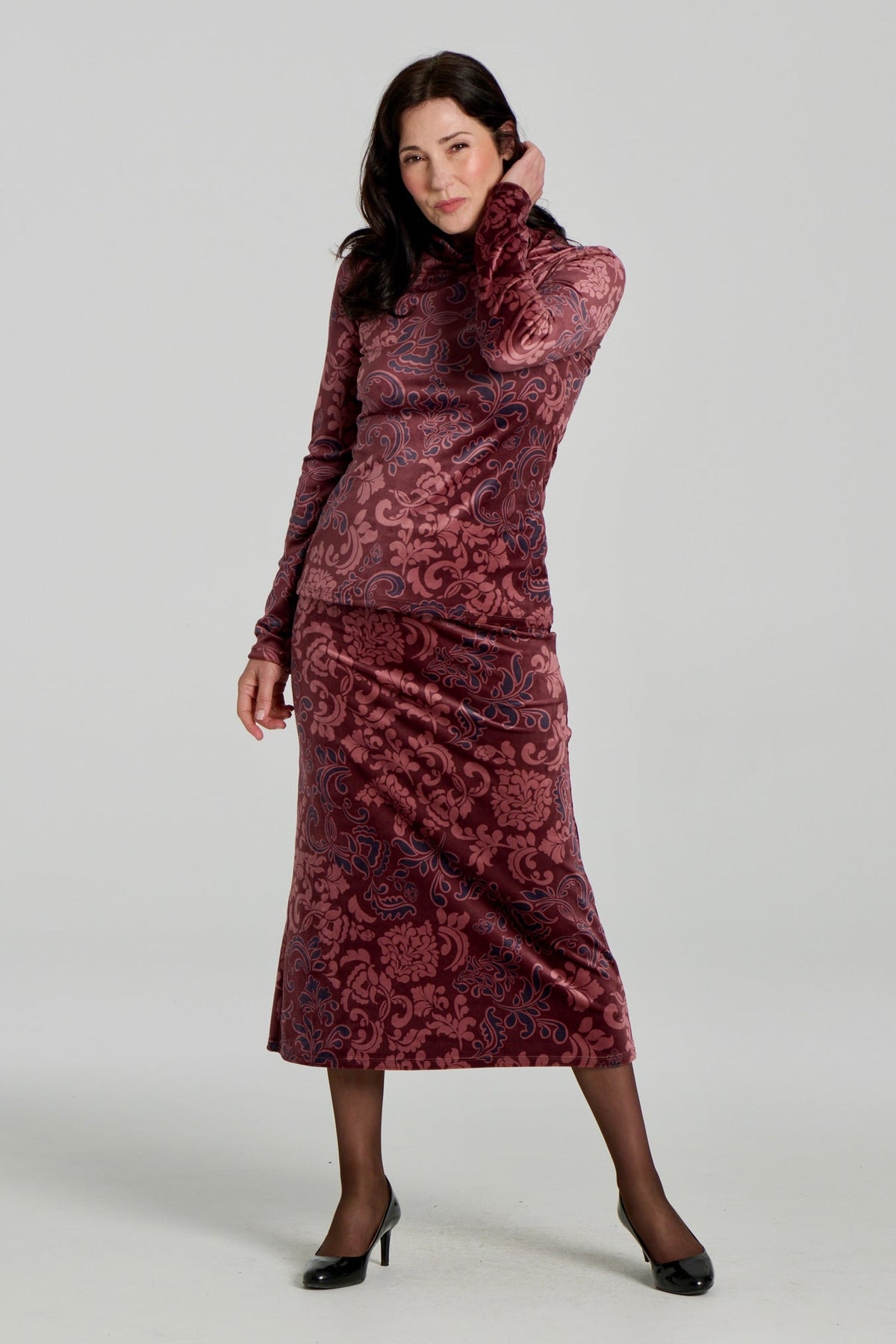 A woman wearing the Everdeen Sweater by Kollontai, a velvet turtleneck in Burgundy paisley print. She is wearing it with the matching Winslet Skirt, a 7/8 A-line pull-on skirt, and standing in front of a white background