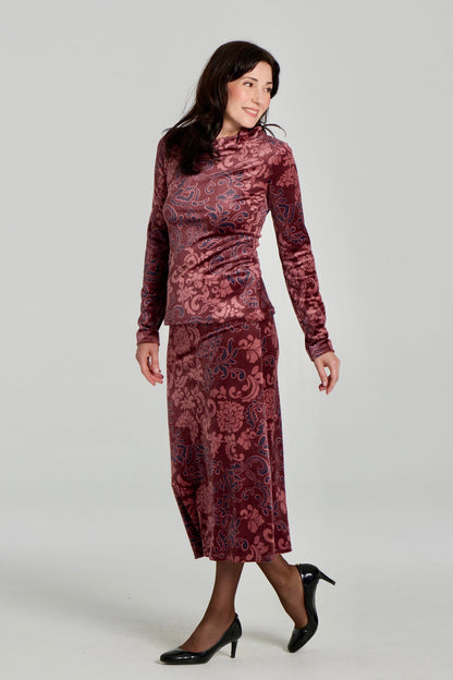 A woman wearing the Everdeen Sweater by Kollontai, a velvet turtleneck in Burgundy paisley print. She is wearing it with the matching Winslet Skirt, a 7/8 A-line pull-on skirt, and standing in front of a white background