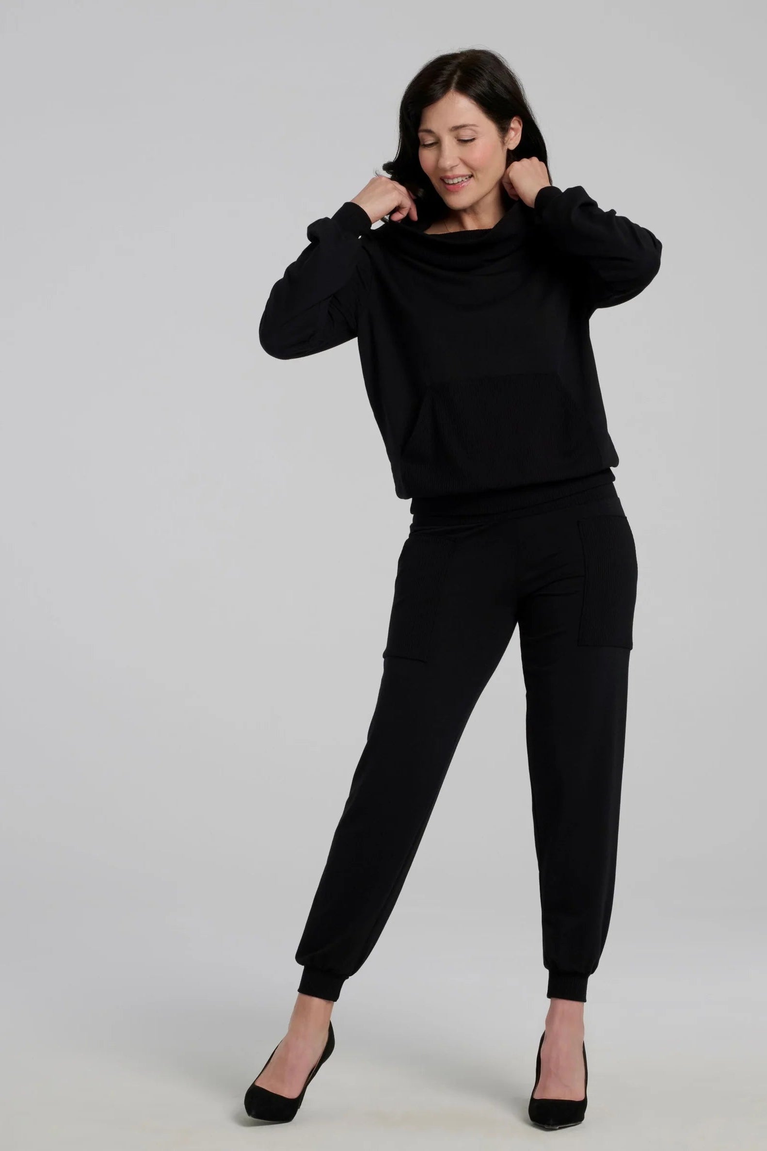 A woman wearing the Jullian Sweater by Kollontai in Black, with a cowl neck, long sleeves, and a kangaroo pocket. She is wearing it with black pants and standing in front of a white background. 