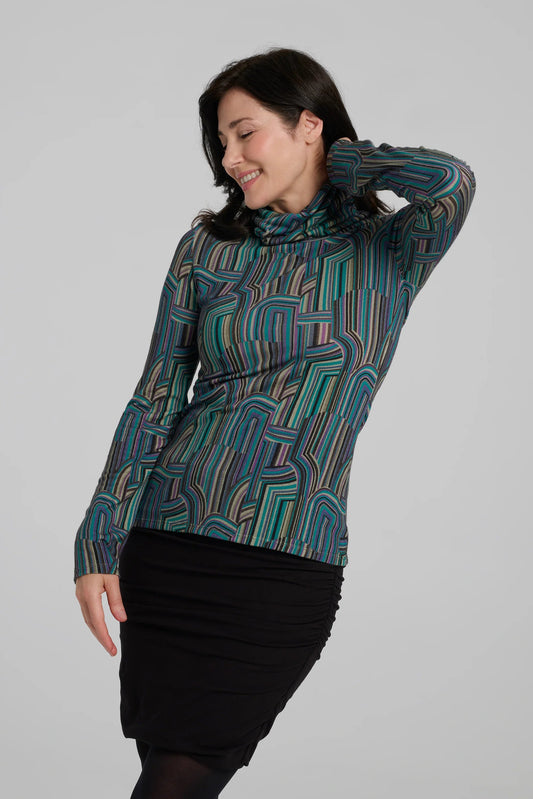 A woman wearing the Yrene Top by Kollontai in Turquoise, a geometric print turtleneck. She is wearing it with a black skirt and standing in front of a grey background. 