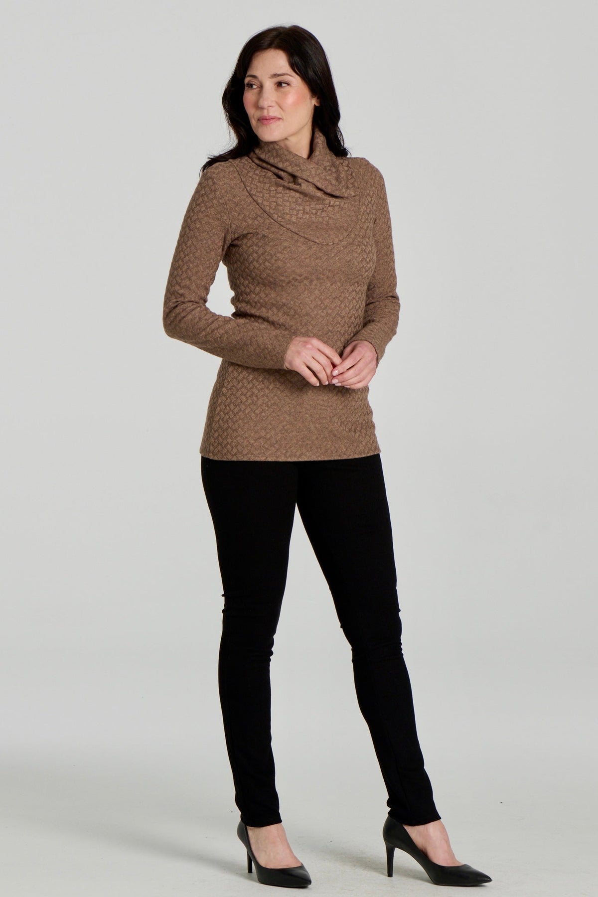 A woman wearing the Eloi Sweater by Kollontai in Brown, a slim fitting textured sweater with a large crossover collar. she is wearing it with black pants and standing in front of a white background. 